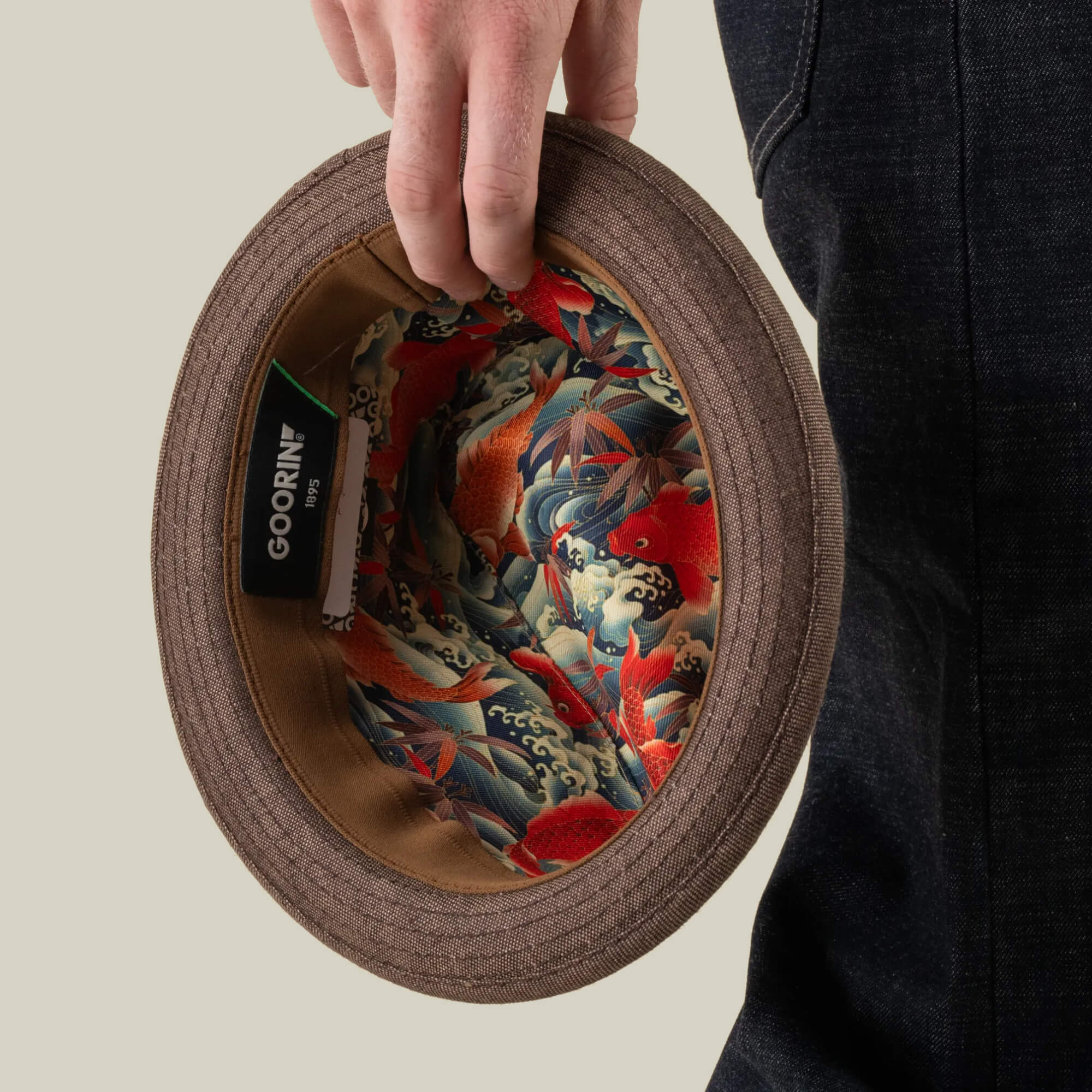 A person holds the brown Charlie Pants by Goorin Bros., showcasing an inner lining adorned with a vibrant koi fish and wave pattern—a striking detail that reflects Goorin Bros.’s signature craftsmanship and attention to detail.