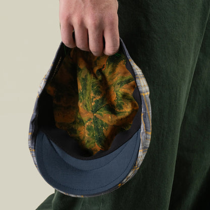 A person holding the Point Lobos 6 Panel Ivy cap by Goorin Bros. by its brim displays the inner lining, which features a green and orange leaf pattern, against a light background. Handmade in the USA from a breathable cotton blend, this blue plaid cap offers all-day comfort and style.