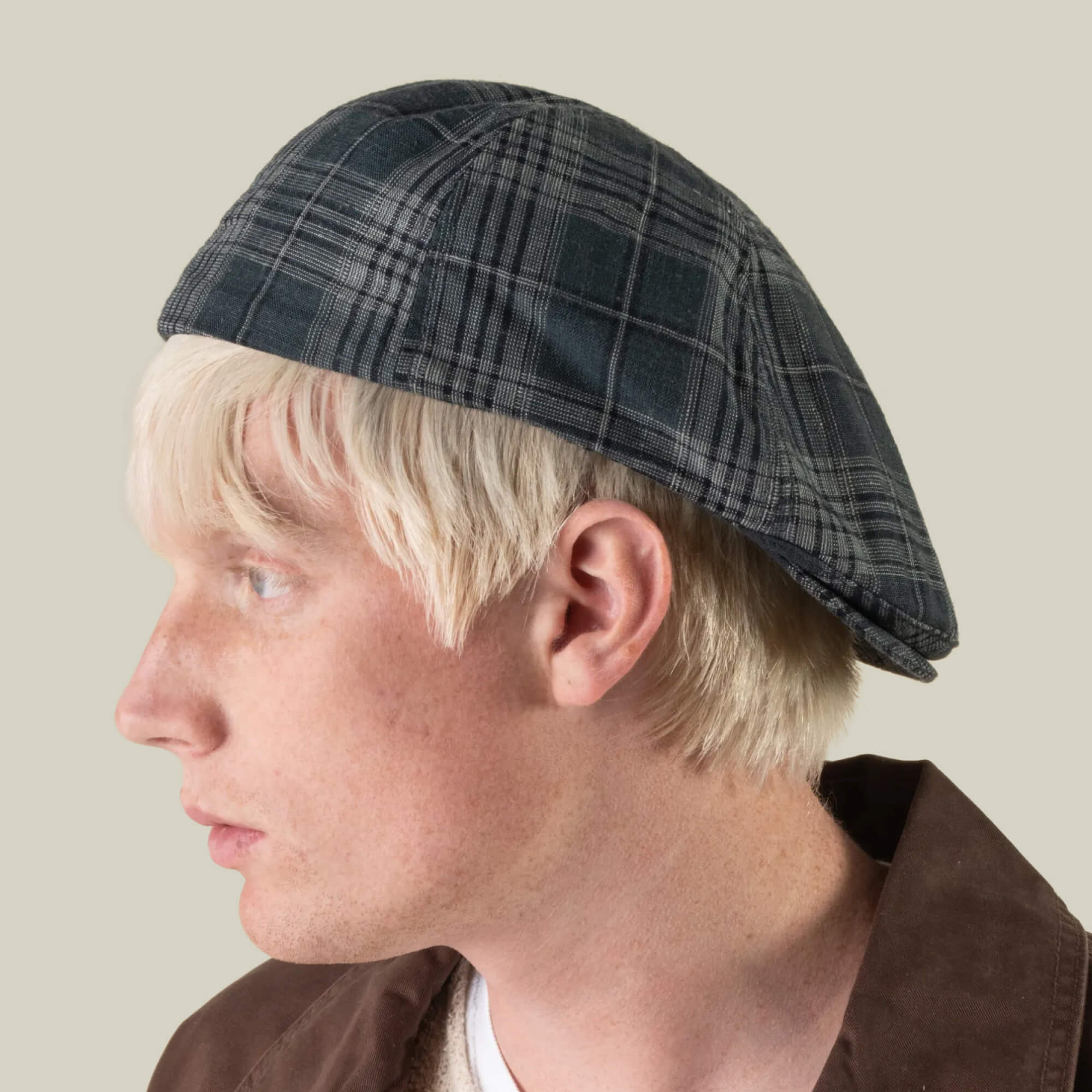 A person with blond hair, seen from the side against a plain light background, is wearing the Goorin Bros. Glenwood Grove flat cap—a gray plaid style hand-cut in the USA from 100% cotton.