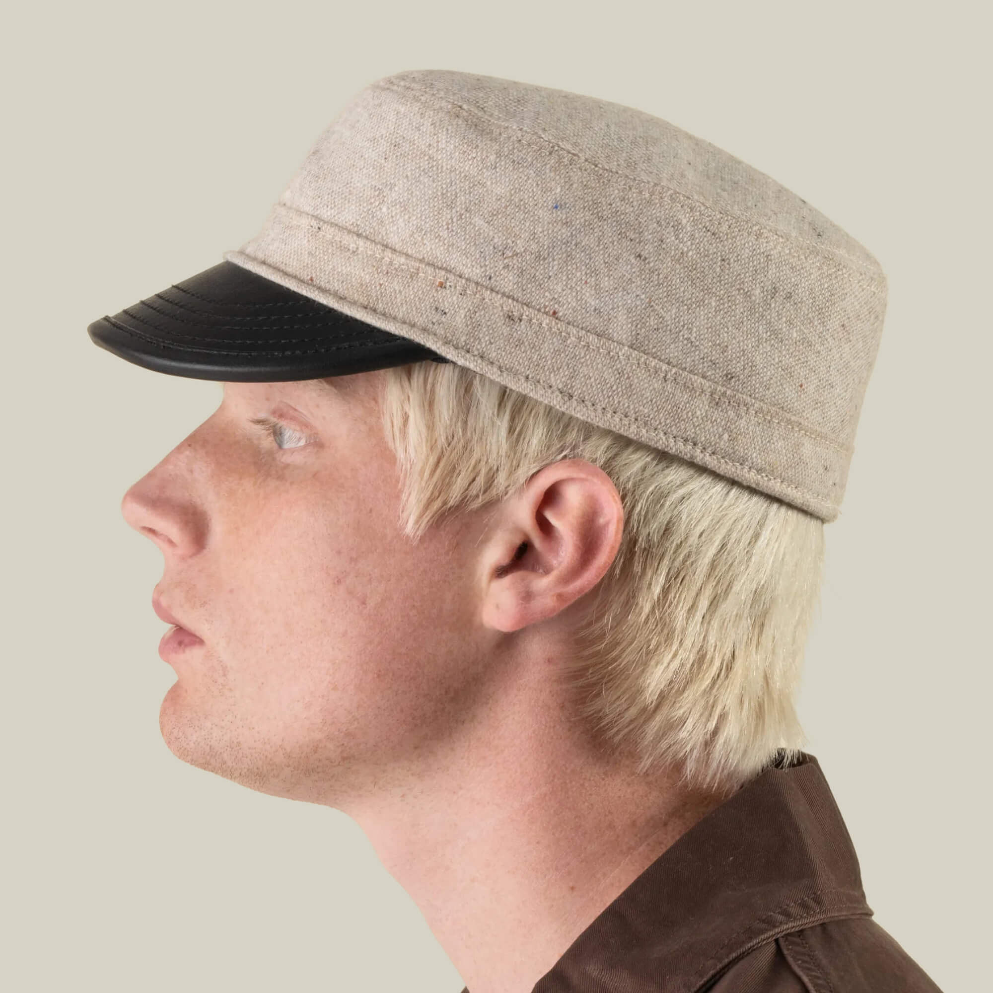 A person in profile wears the South Boulder cap by Goorin Bros.—a light beige wool and leather cadet cap with a black visor—paired with a brown shirt against a plain light background; each hat is hand cut and sewn in the USA for the heritage-minded.