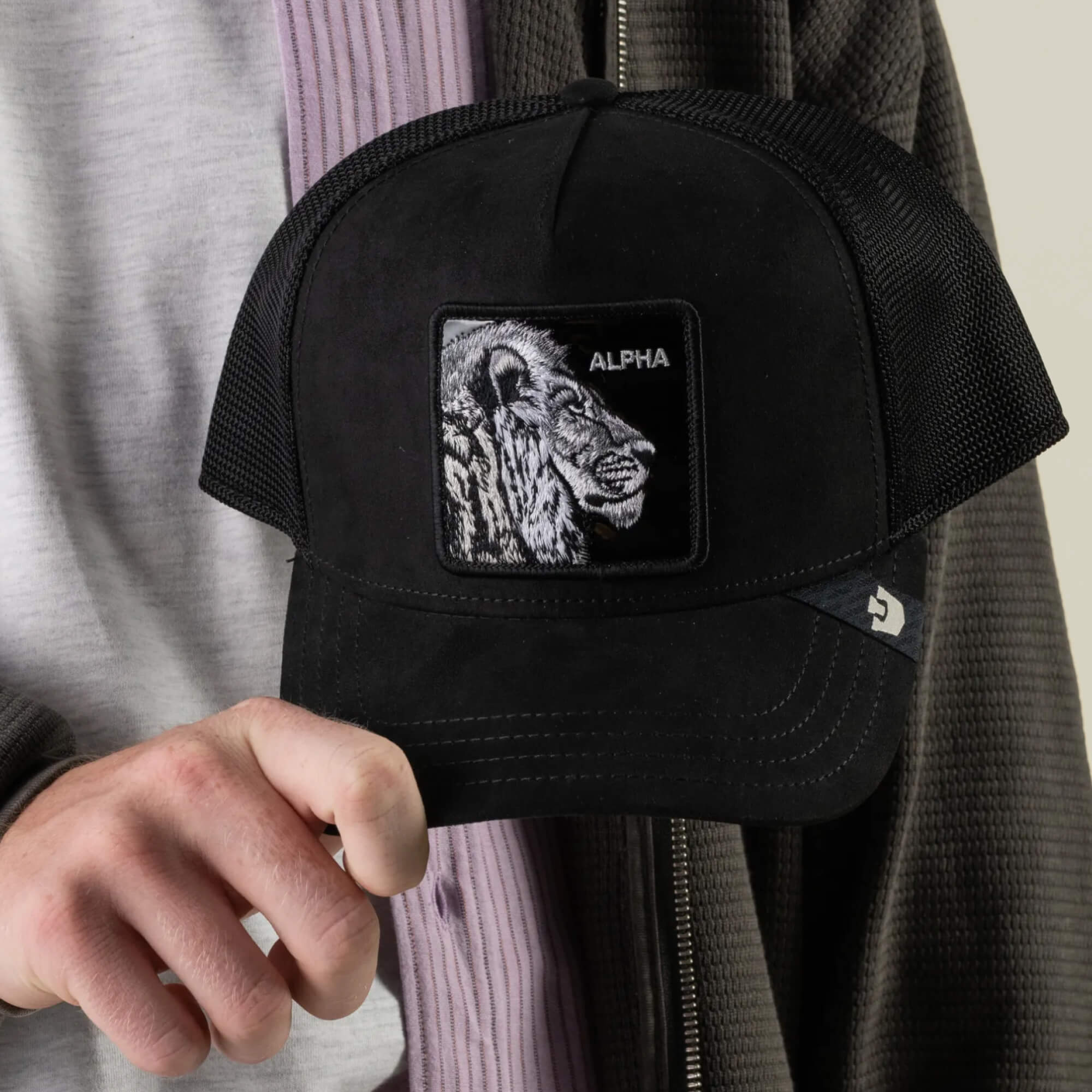 A person holds the Goorin Bros. Black Out Lion trucker hat, which features a black mesh design and a patch with an embroidered lion and the word “ALPHA” above it.