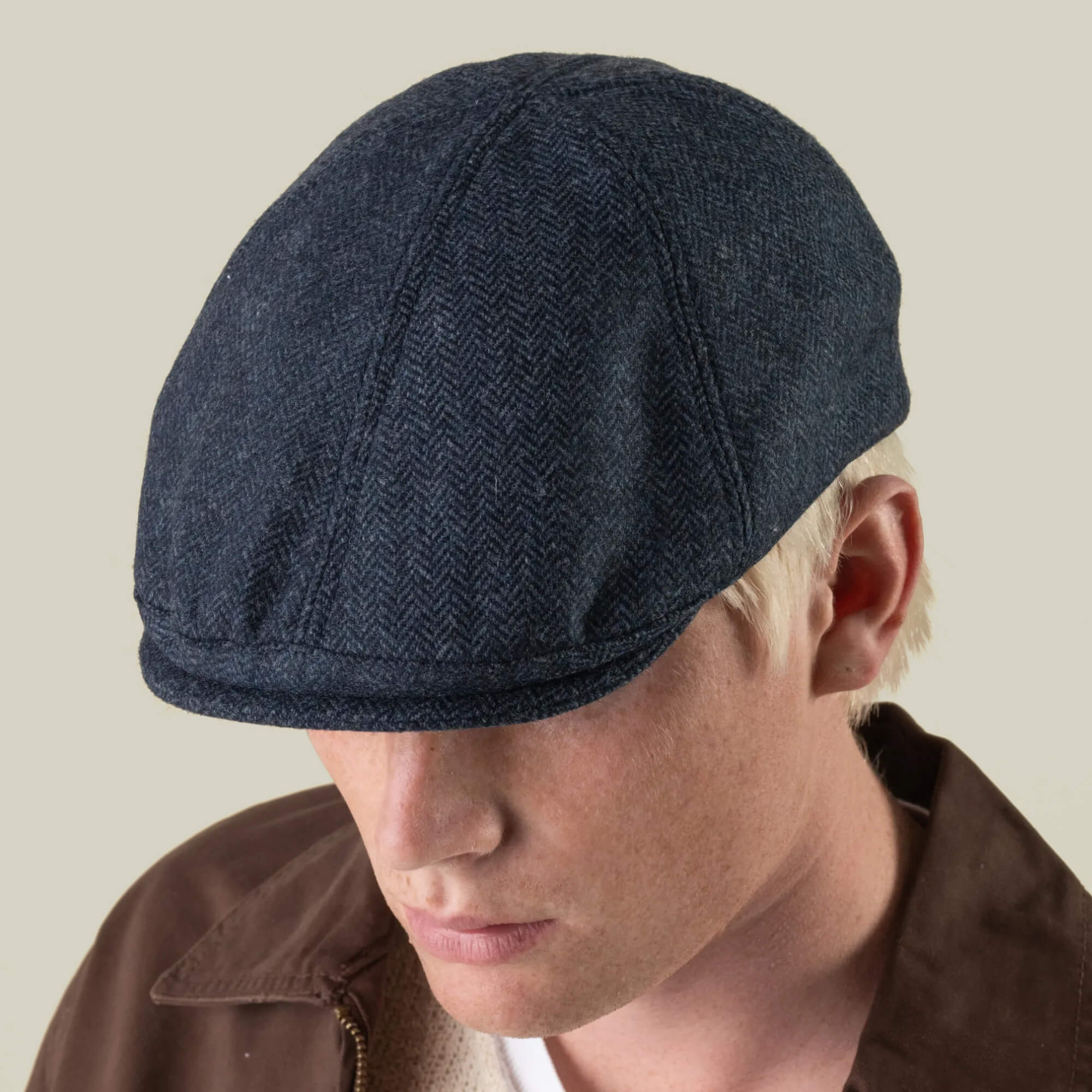 A person with light skin and blond hair wears the Harry Bones flat cap by Goorin Bros.—a dark blue cap crafted from 100% wool and made in the USA—paired with a brown jacket, facing slightly downward against a neutral background.