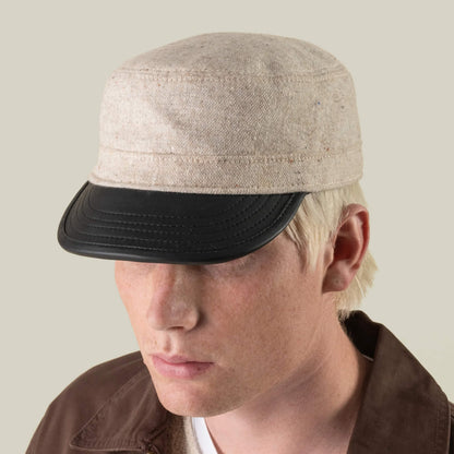 A person wears the South Boulder cadet cap by Goorin Bros.—heritage-inspired, hand-cut and sewn in the USA, featuring a black leather brim—paired with a brown jacket and a neutral expression against a plain background.