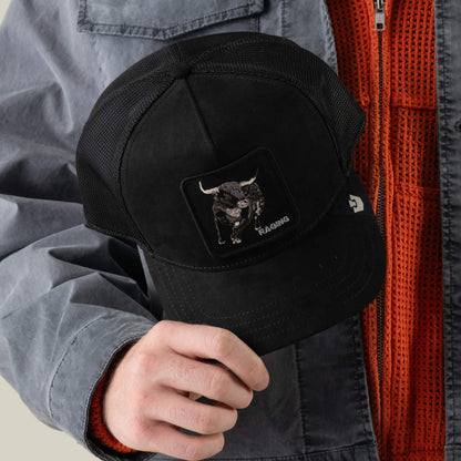A person in a gray jacket and orange textured shirt holds a black polyester mesh cap from Goorin Bros., featuring a “Suede Bull” patch.