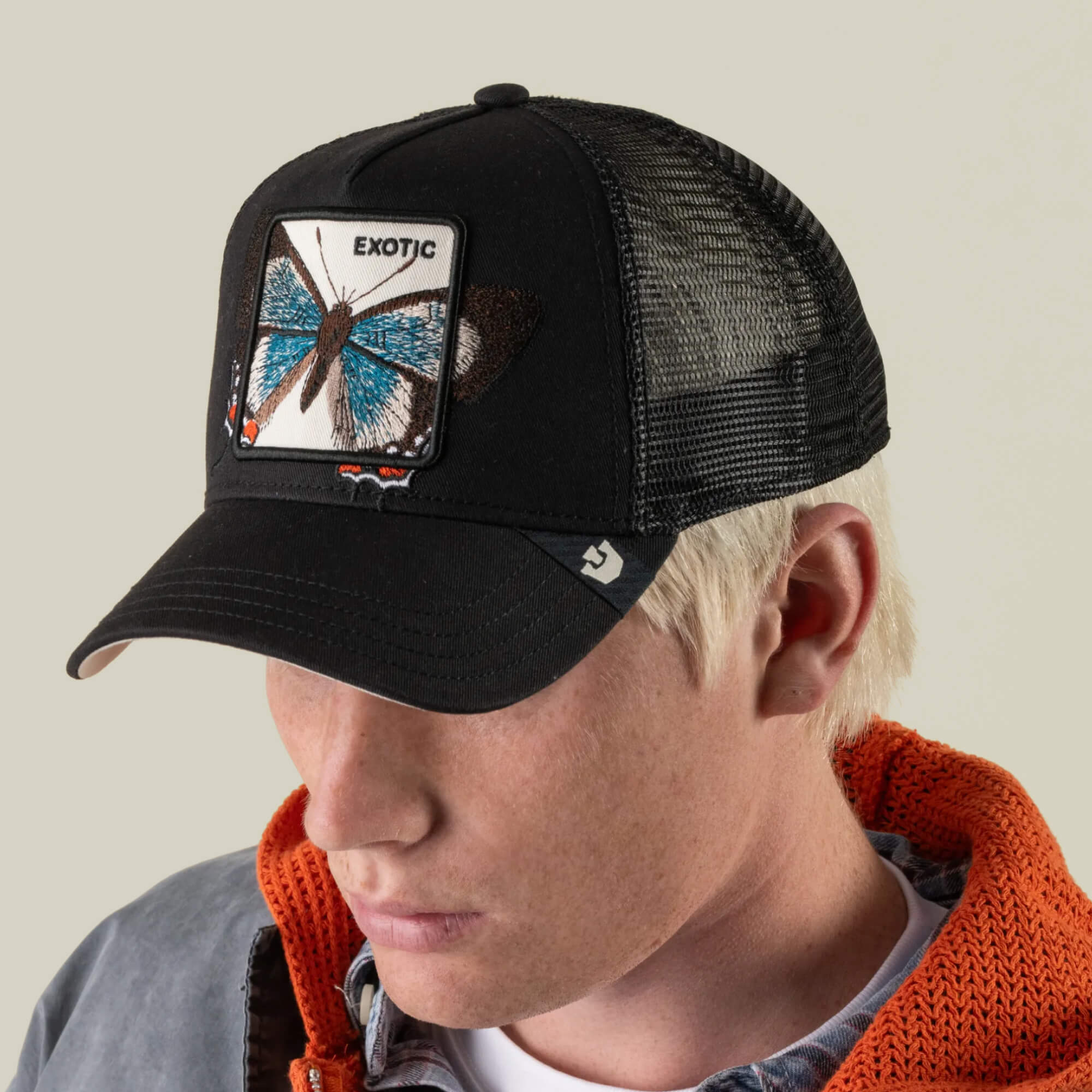 A person wearing the Goorin Bros. Over the Line Butterfly black breathable mesh cap, featuring a patch with a blue butterfly and the word “EXOTIC,” paired with an orange hoodie and gray jacket.