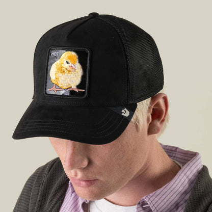 A person wearing the Goorin Bros. “Black Out Chick” trucker baseball cap, which features a black mesh design and a yellow chick patch labeled “HOT,” paired with a casual button-up shirt and jacket.