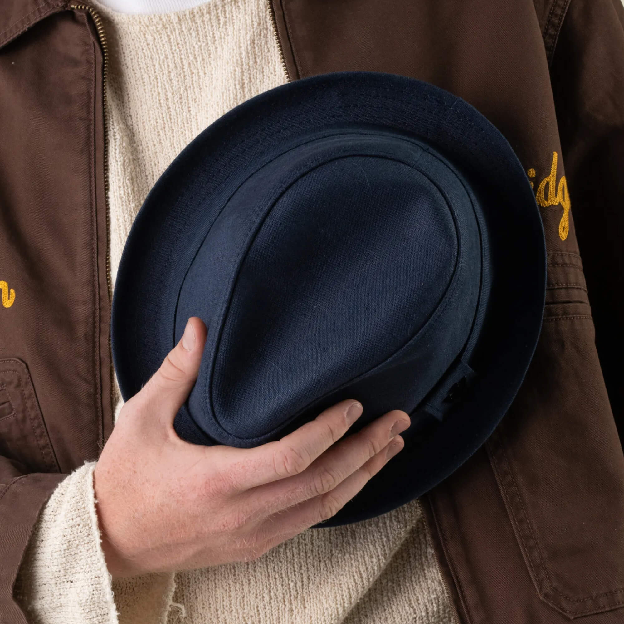 A person wearing a brown jacket and cream sweater holds the Goorin Bros. Charlie Pants, hand-cut in the USA, against their chest with one hand.