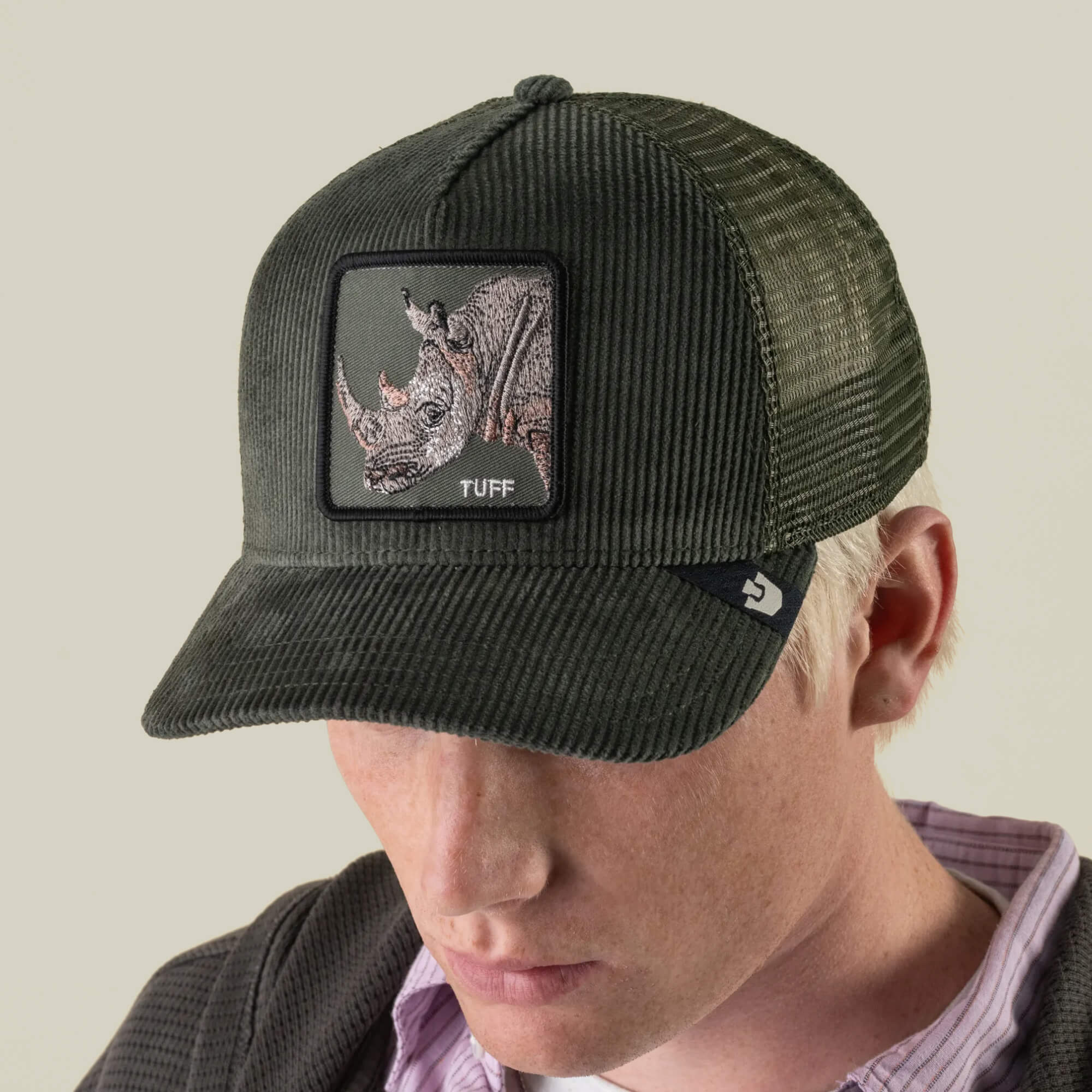 A person wearing the Goorin Bros. Corduroy Tuff hat—a dark green trucker hat crafted from a durable polyester-cotton corduroy blend, featuring a rhino embroidered patch with the word “TUFF”—looks downward against a plain background.