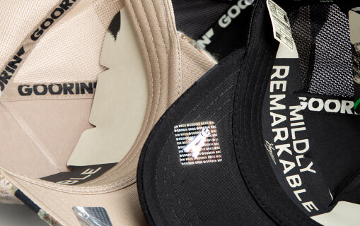 Close-up view of the insides of two Goorin trucker hats; one tan and one black. The black cap has MILDLY REMARKABLE printed on the inner brim, and both have visible brand labels and stickers.
