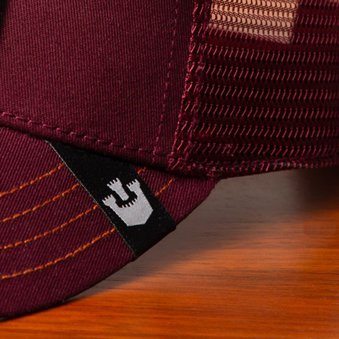 Close-up of a maroon mesh trucker hat by Goorin on a wooden surface, showing a black and white woven logo tag on the brim—perfect for fans of bold hat designs.