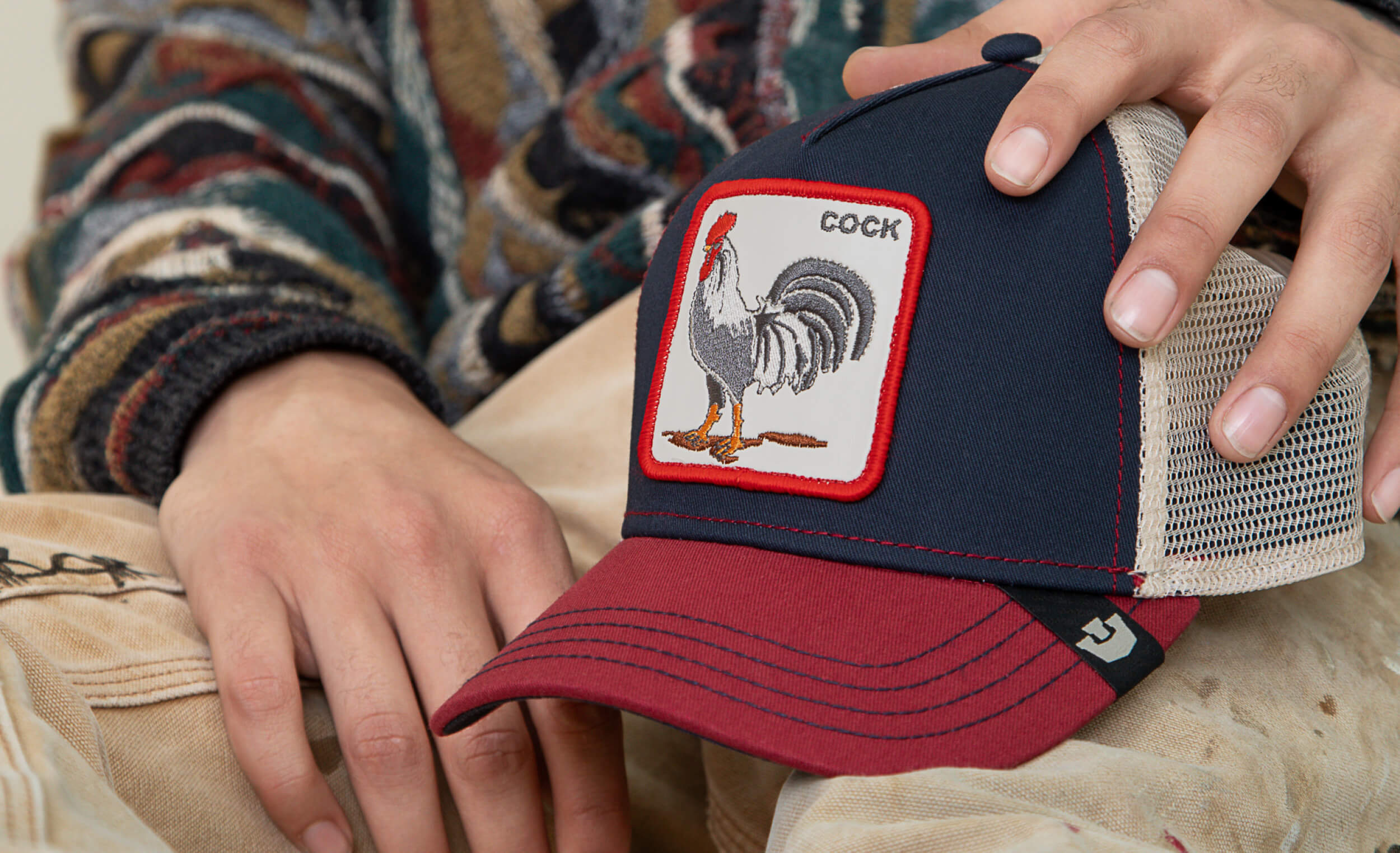 A person holds a navy blue and red trucker hat with a rooster patch labeled COCK on the front, showcasing one of the bold hat designs that set trucker hats apart.