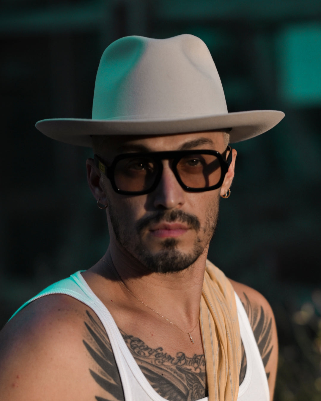 A man wearing a light-colored Goorin fedora hat, large black sunglasses, a white tank top, and a yellow scarf draped over his shoulder, with visible chest tattoos.