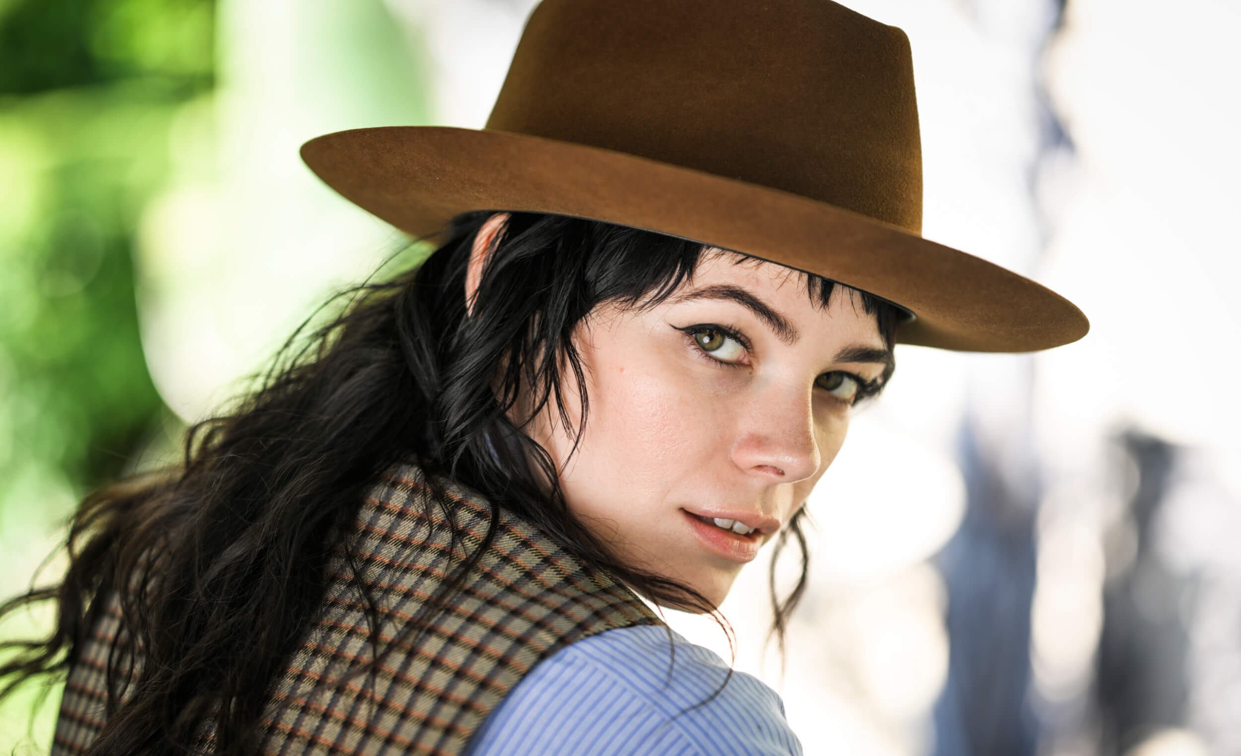 A person with long dark hair, wearing a brown Goorin trucker hat and plaid vest, looks over their shoulder with a neutral expression.