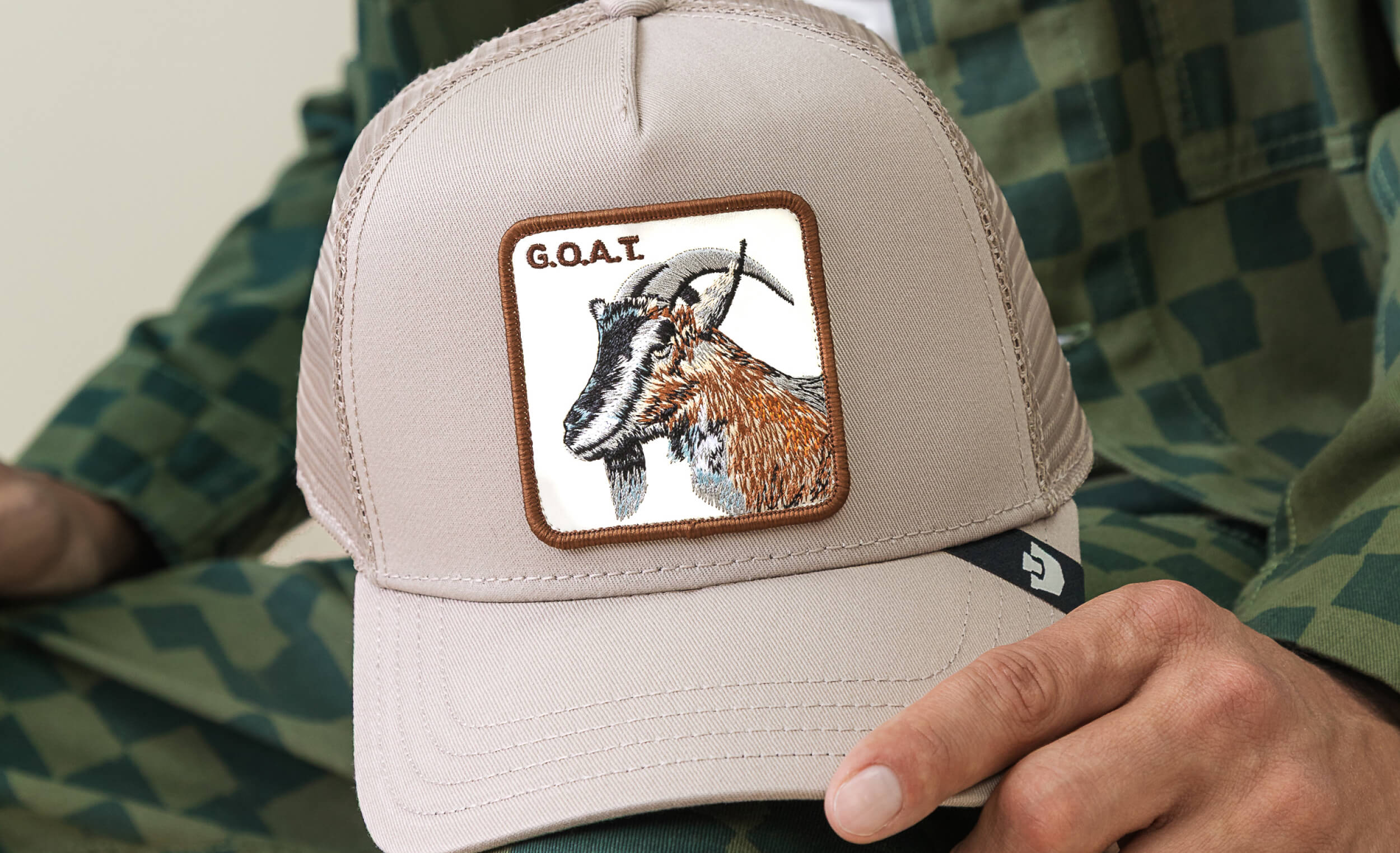 Beige trucker hat with a goat patch and 'G.O.A.T.' text, worn by a person in camouflage clothing.