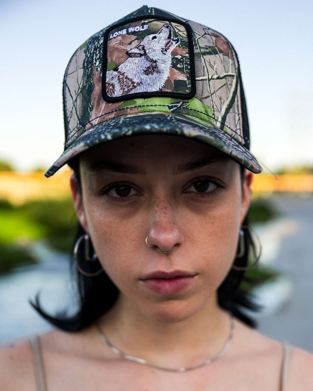 Person with hoop earrings and a nose ring wears an American made camouflage cap featuring a wolf and the text Lone Wolf. The background is an outdoor setting out of focus.
