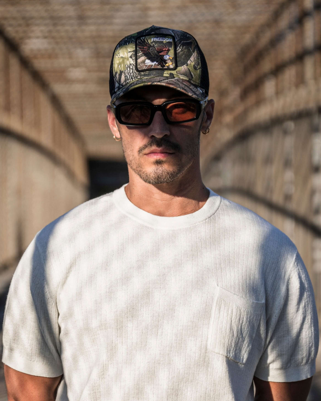 Man wearing a white short-sleeve shirt, dark sunglasses, and a camouflage cap stands on a bridge with metal fencing in the background, showcasing American made durable hats perfect for any outdoor adventure.