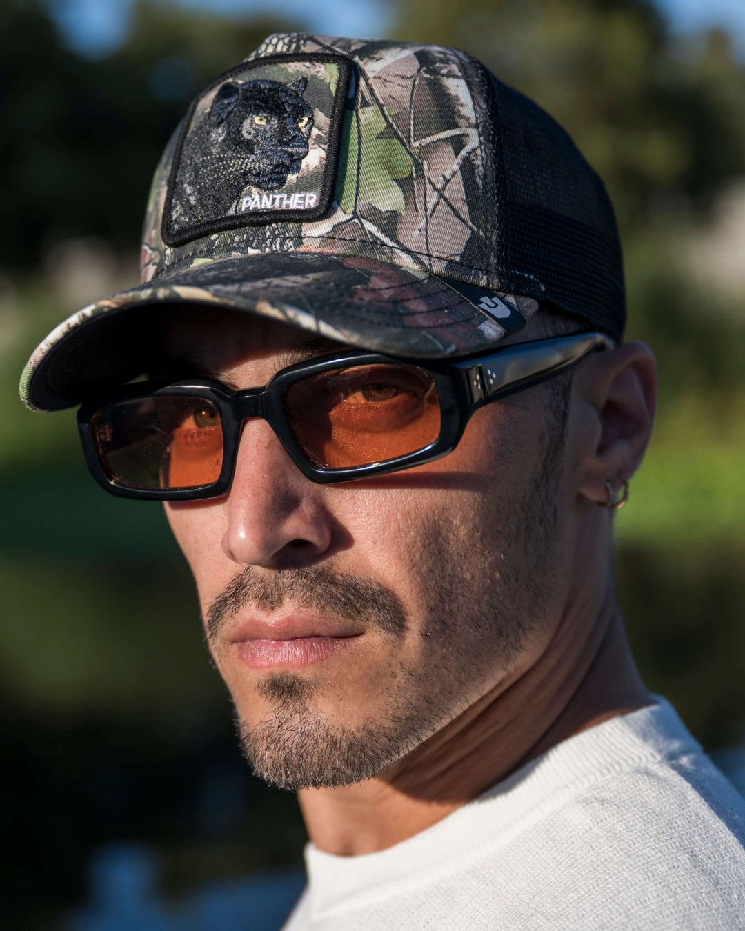 Man wearing a camo cap with a Goorin panther patch, sunglasses with orange lenses, a small hoop earring, and a white shirt, outdoors in sunlight. A perfect blend of American made style beyond classic fedora hats.