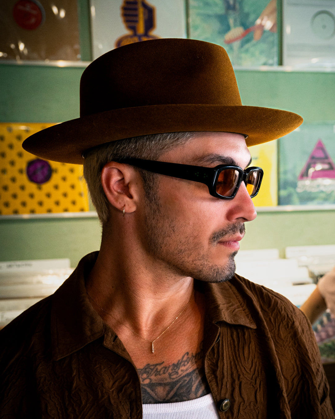 A man with short blonde hair, wearing a bold Goorin hat, dark sunglasses, a brown shirt, and a thin necklace stands in front of a wall with records.