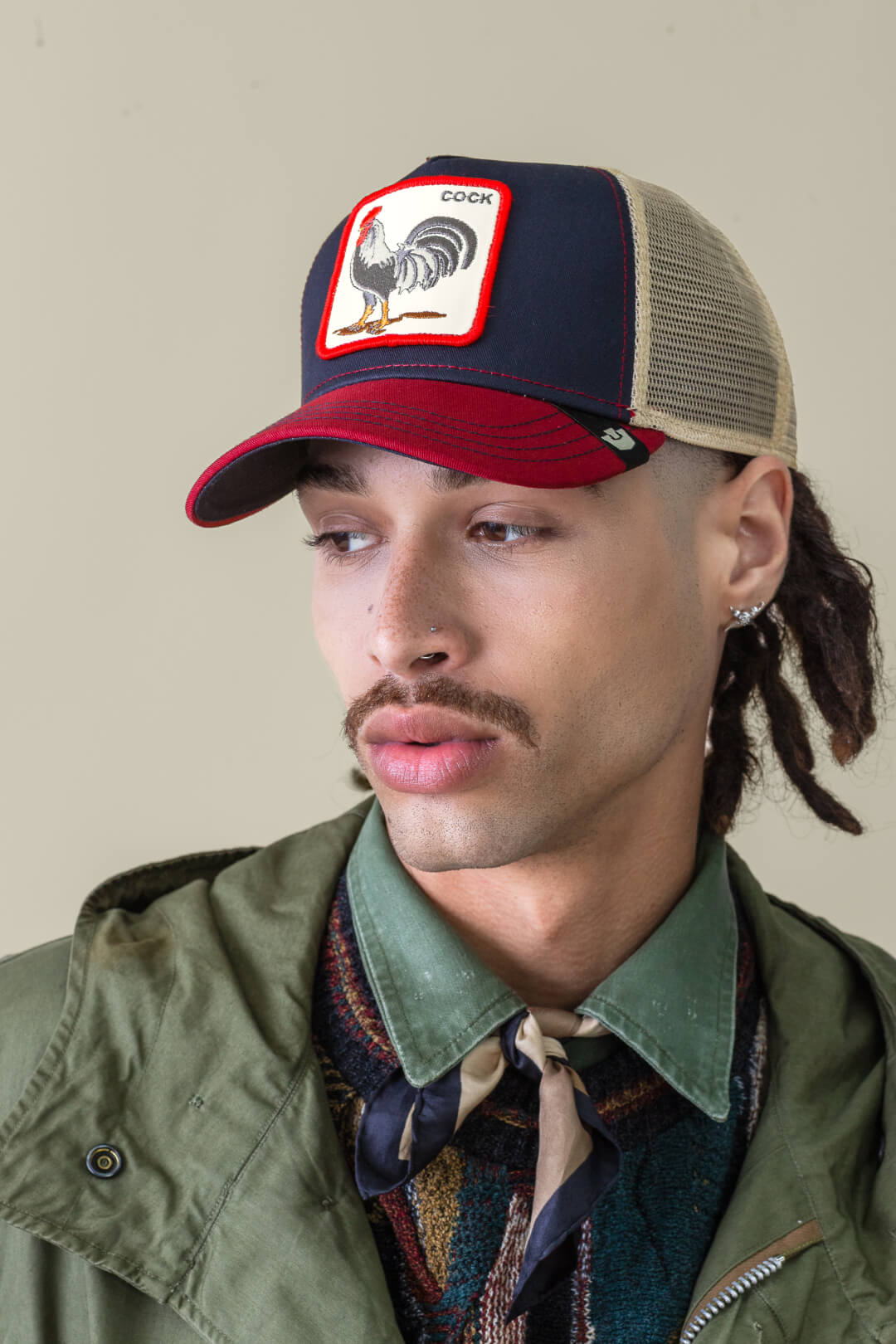 Person wearing an American made mesh baseball cap with a rooster patch, green jacket, patterned sweater, collared shirt, and scarf, looking to the side.