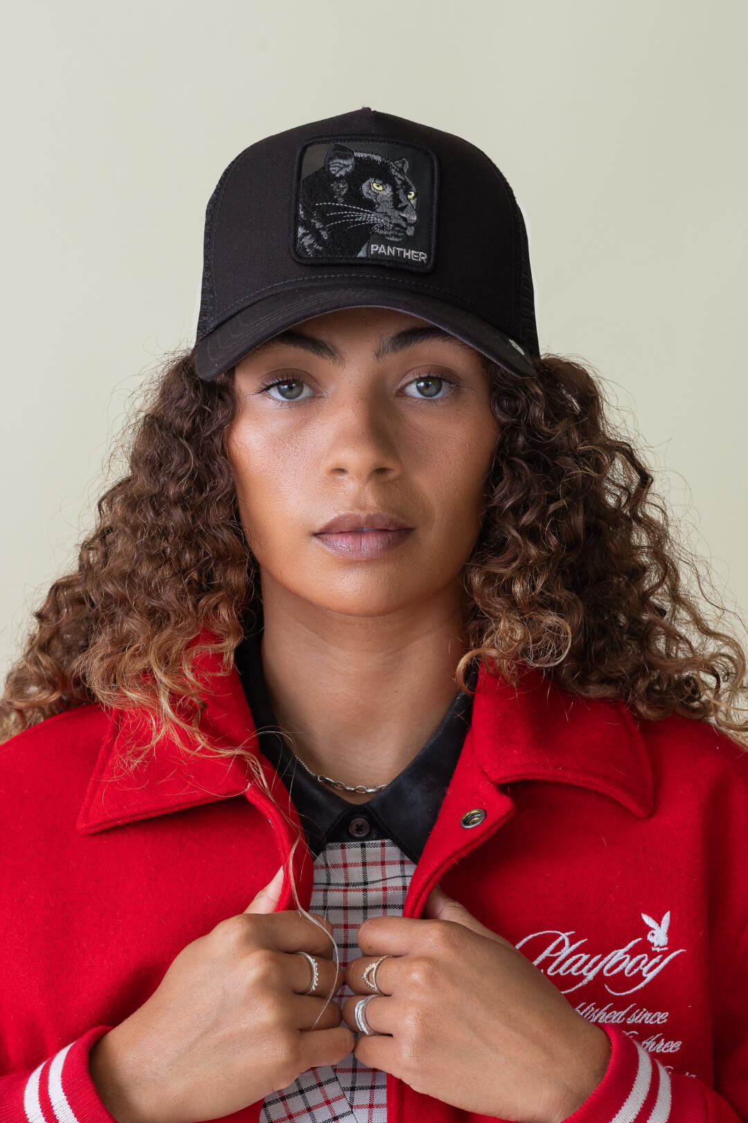 A woman with curly hair wears a black Goorin trucker hat featuring a panther patch and a red jacket over a plaid shirt, looking directly at the camera.