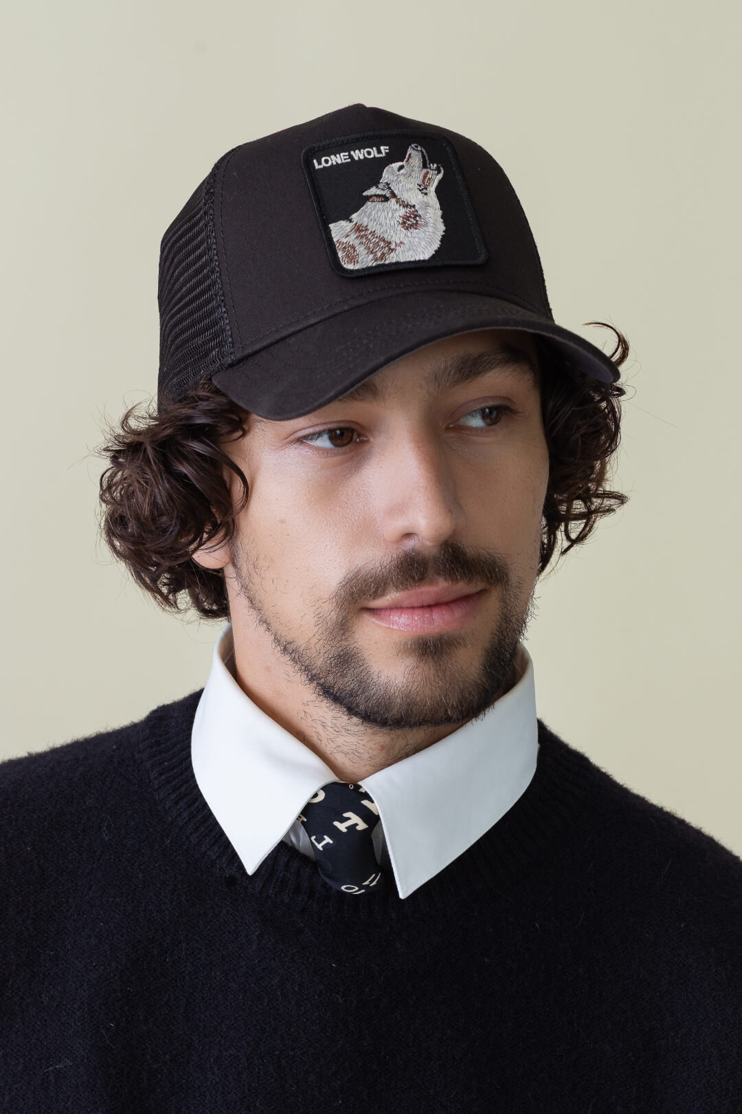 A man with curly hair and facial hair wears an American made black trucker hat featuring a Lone Wolf patch, a black sweater, white collared shirt, and a patterned tie, against a plain background.