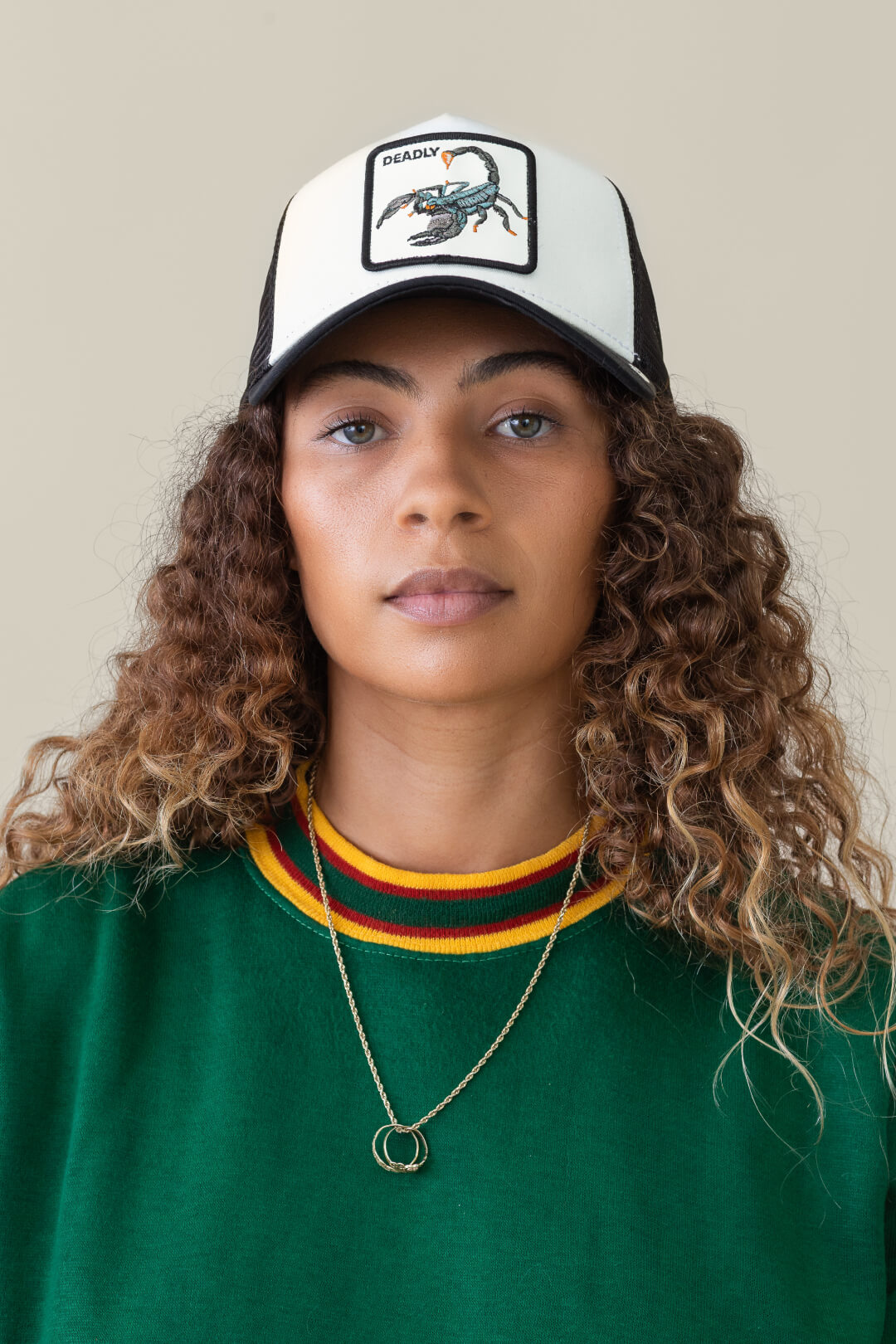 A person with curly hair wearing a green sweater, a gold necklace, and a bold trucker hat featuring a scorpion patch labeled Deadly, standing against a plain background.