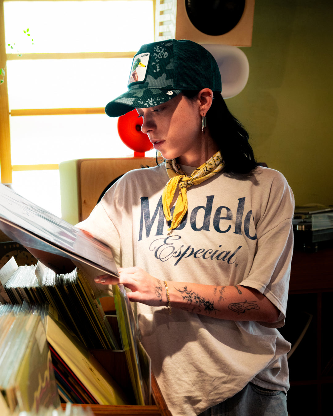 A person wearing a Modelo Especial t-shirt and a durable green trucker hat browses through vinyl records in a well-lit room.