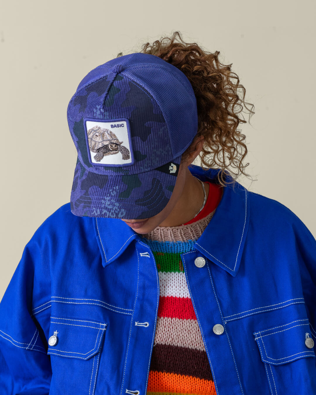 Person wearing an American made blue camouflage-patterned trucker hat with a patch, a blue jacket, and a colorful striped sweater, looking downward against a plain background.