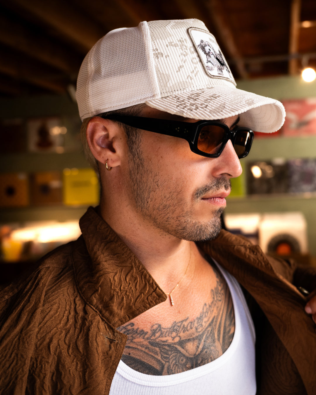 A man wearing a white cap—one of those classic American made trucker hats—black sunglasses, and a brown jacket over a white shirt, with visible tattoos on his chest, stands indoors with blurred records in the background.