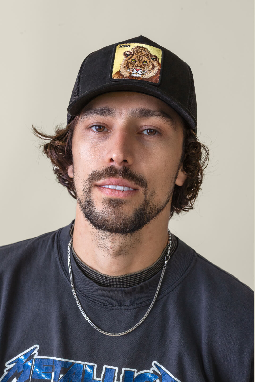 A man with wavy brown hair, a beard, and mustache wears a bold hat design—a black trucker hat with a lion patch—paired with a Metallica t-shirt and silver chain necklace against a plain background.