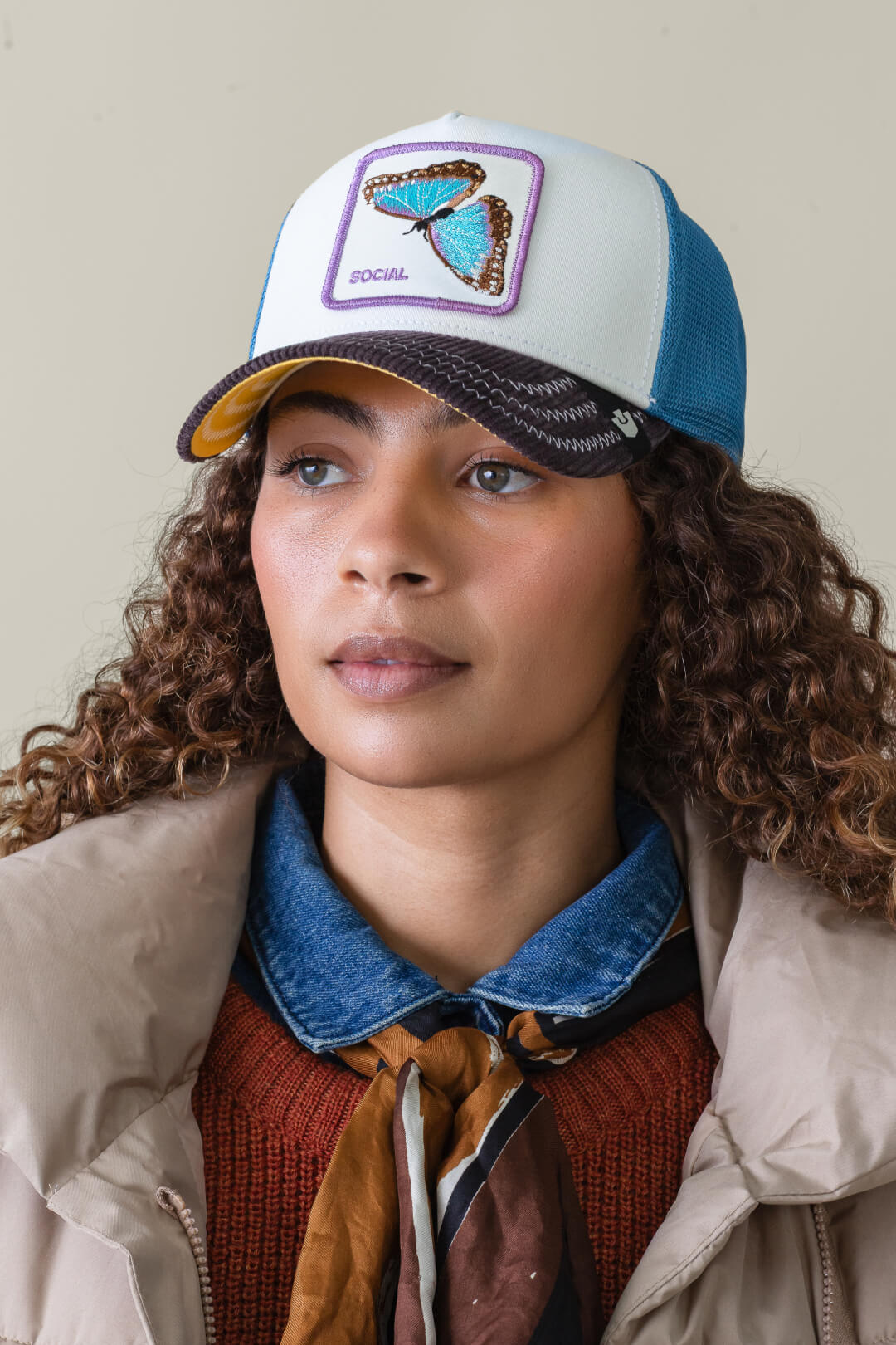 A woman wears a blue and white Goorin cap with a butterfly patch and the word SOCIAL, layered clothing including a scarf, collared denim shirt, and beige jacket—showcasing bold hat designs.