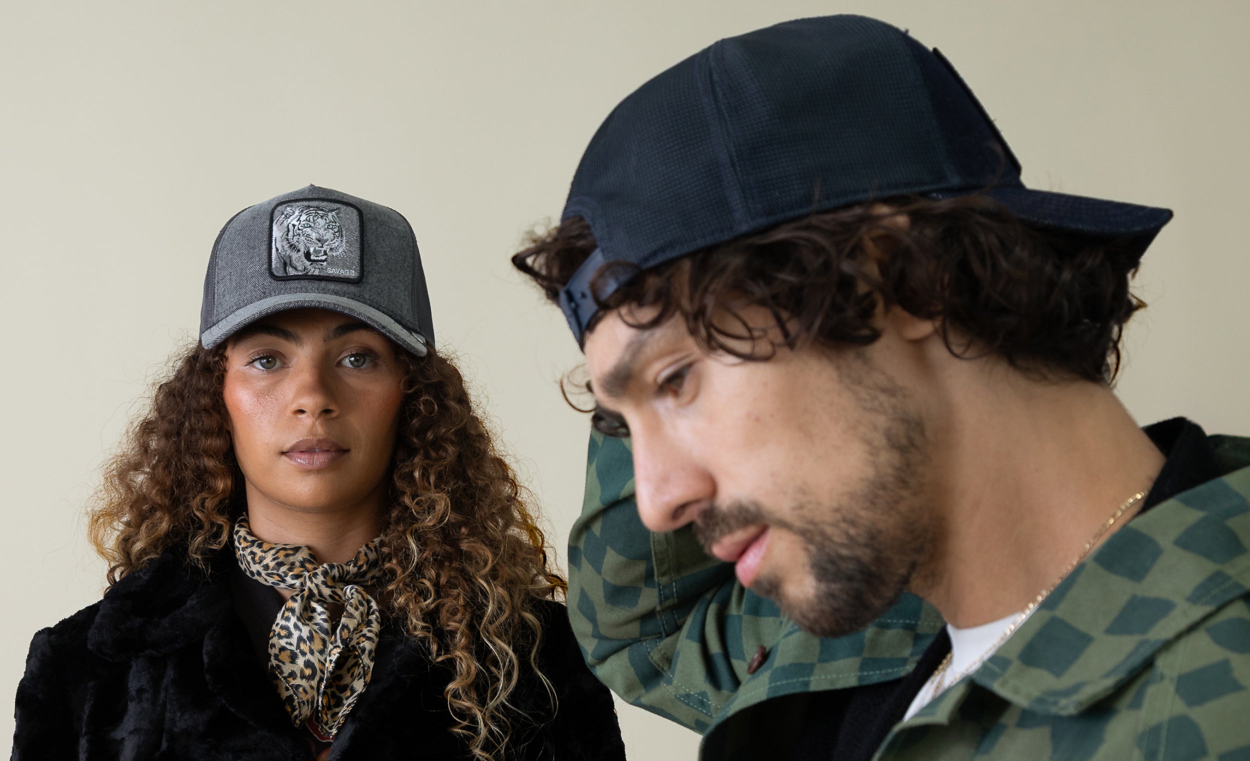 A woman and a man wearing trucker hats; the woman looks at the camera while the man, dressed in a green checkered shirt, looks downward and adjusts his American made Goorin cap.