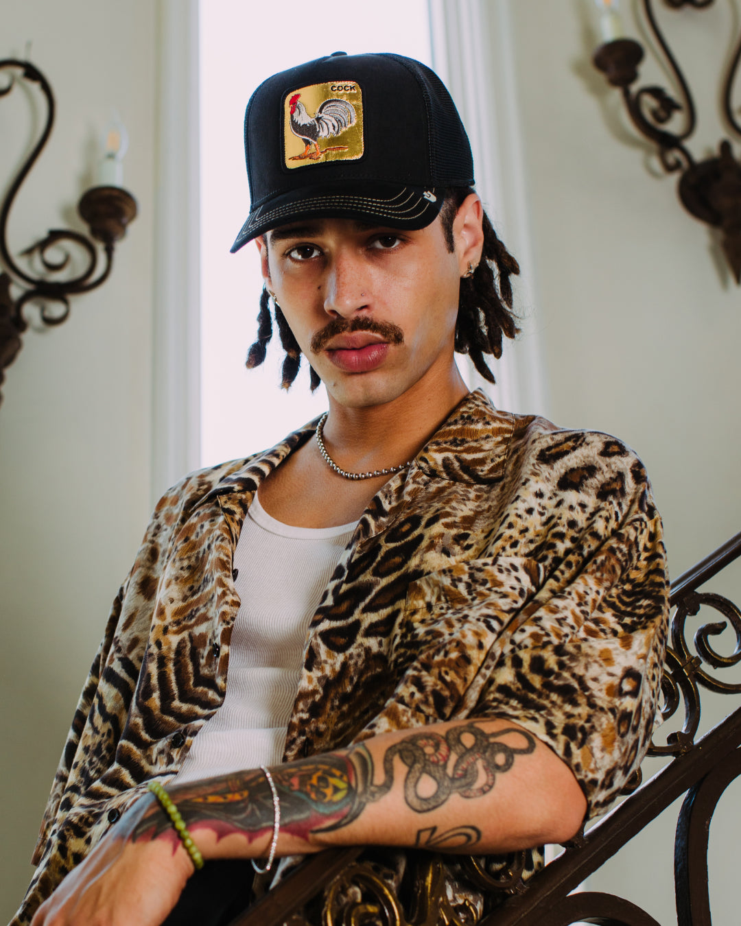 A person with a mustache and tattoos, sporting a Goorin rooster patch cap, a leopard print shirt, and a white tank top, stands by a decorative stair railing in front of tall windows—embracing bold hat designs with confidence.