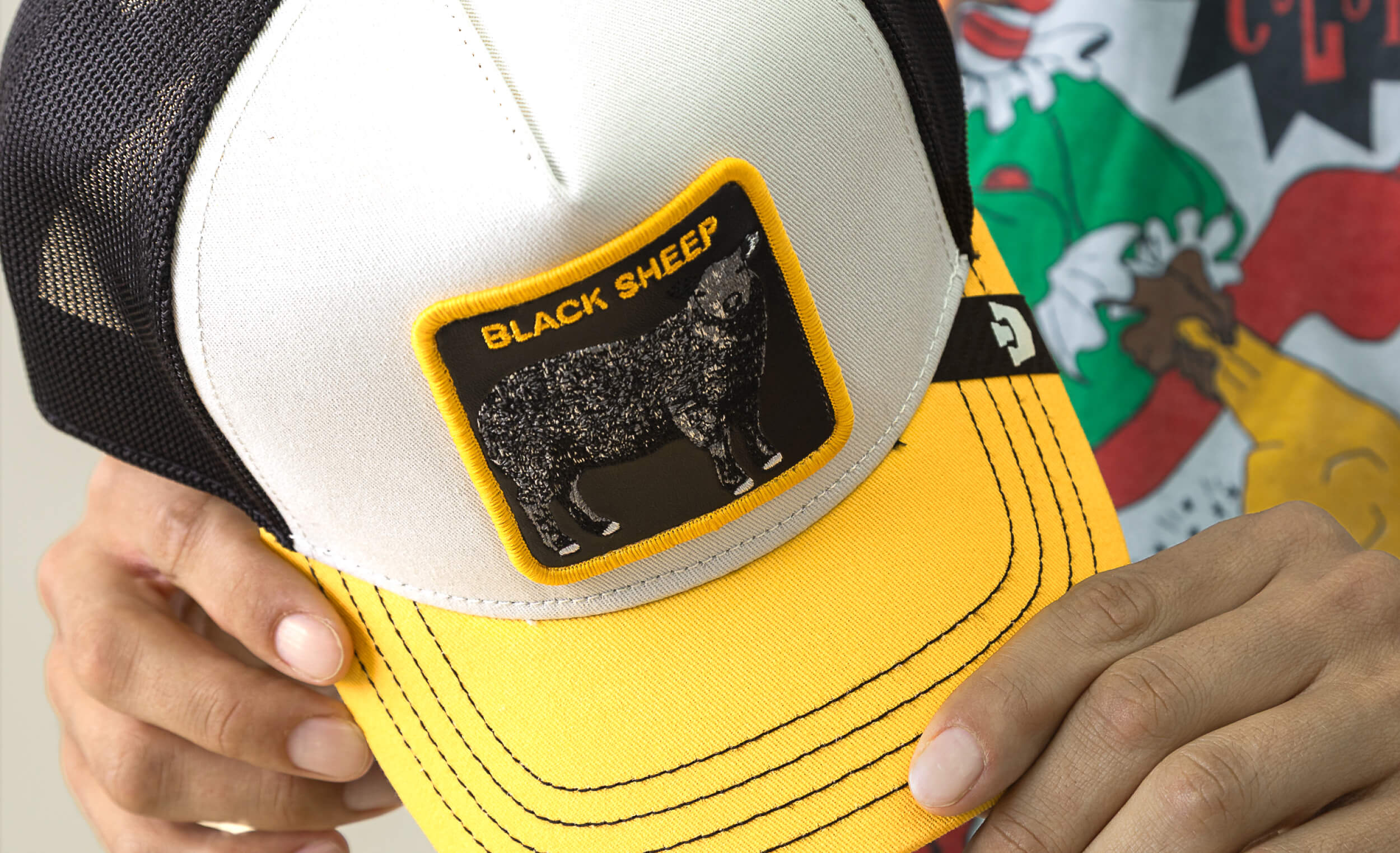 Yellow and black cap with 'Black Sheep' logo held by a person, with a colorful background.