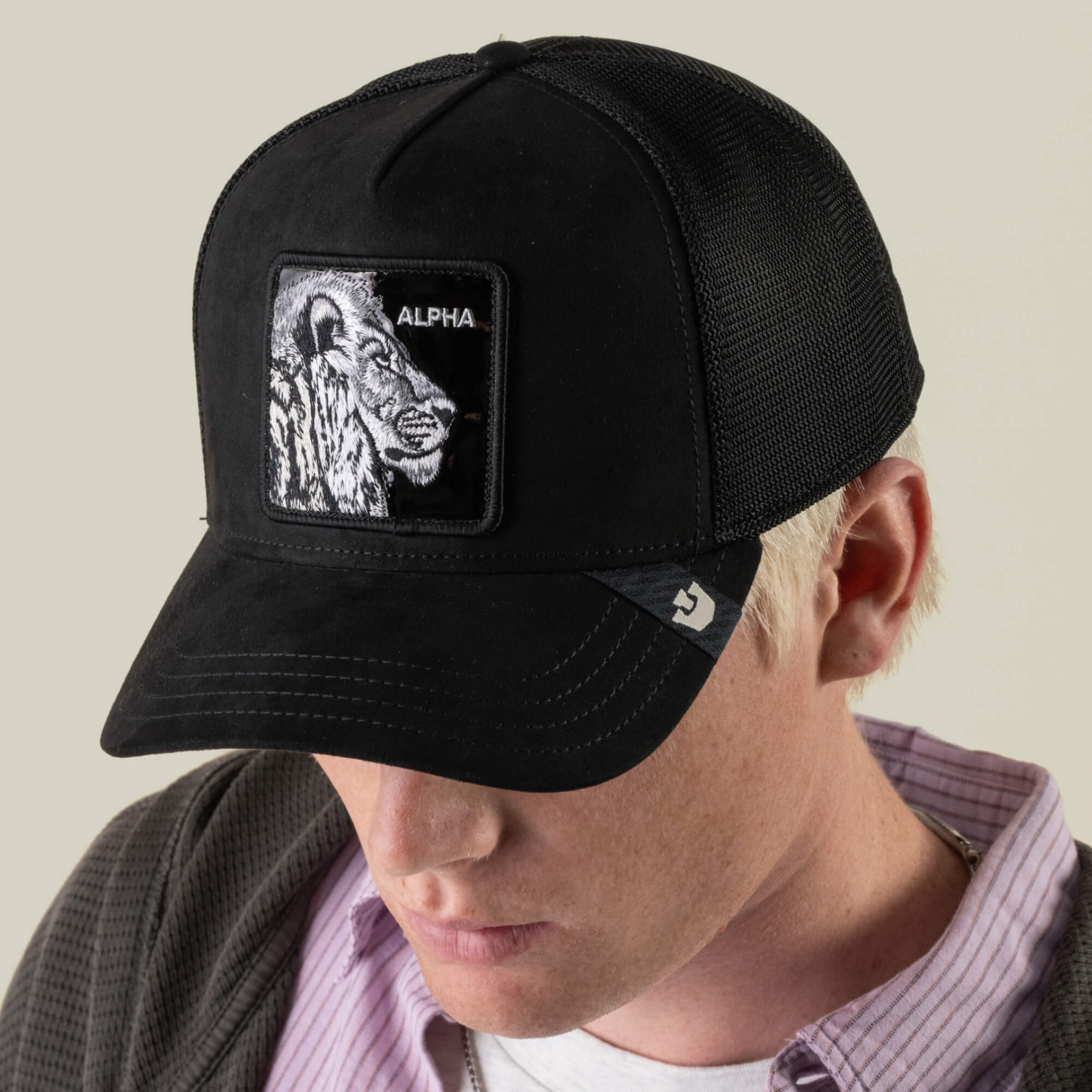 A person with light hair, dressed in a collared shirt and jacket, looks downward while wearing the Goorin Bros. “Black Out Lion” mesh stealth trucker hat, which features an embroidered lion patch labeled ALPHA.