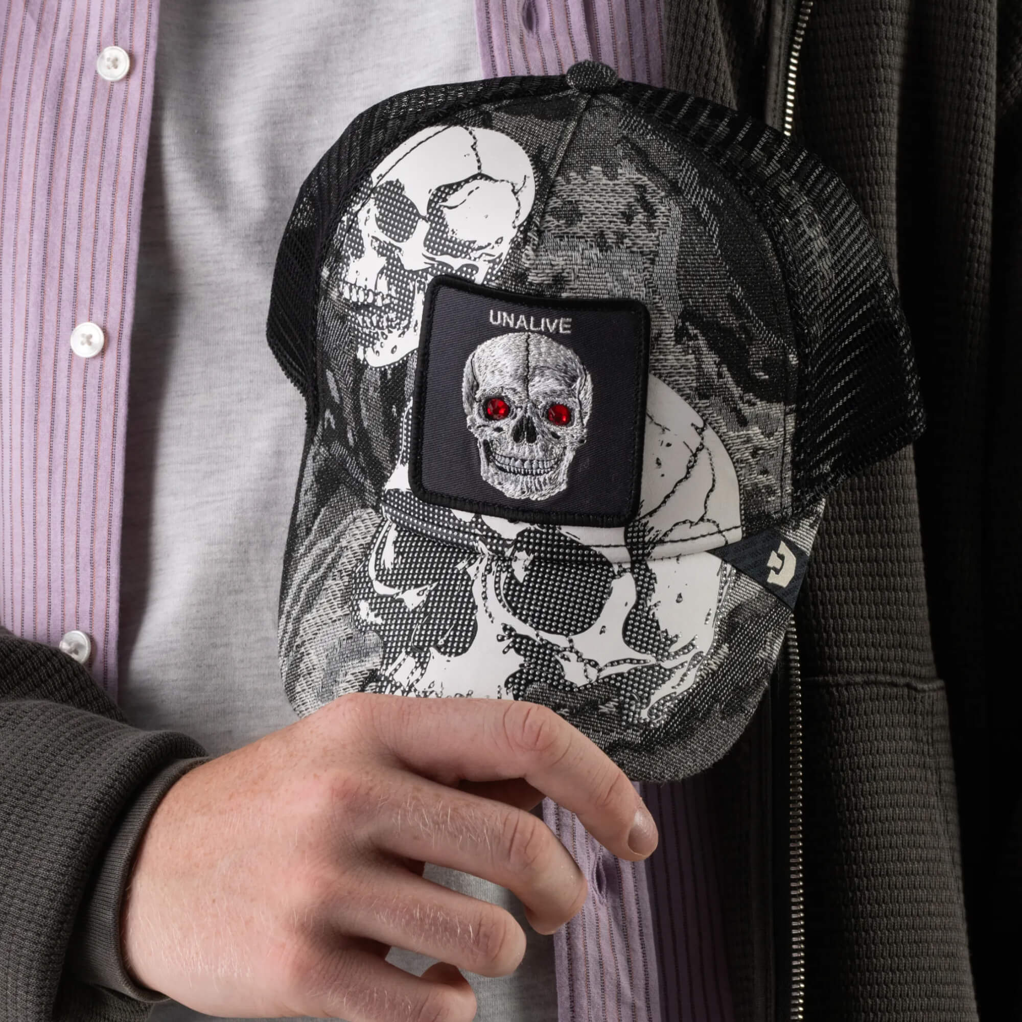 A person holds the Goorin Bros. Bone Head Unalive trucker hat, a black and white cap with a breathable mesh back and striking white skull graphics, including a patch of a skull with red eyes above the word “UNALIVE.” The hat is styled with the individual’s grey shirt and pink striped shirt, echoing classic Goorin Bros. design flair.