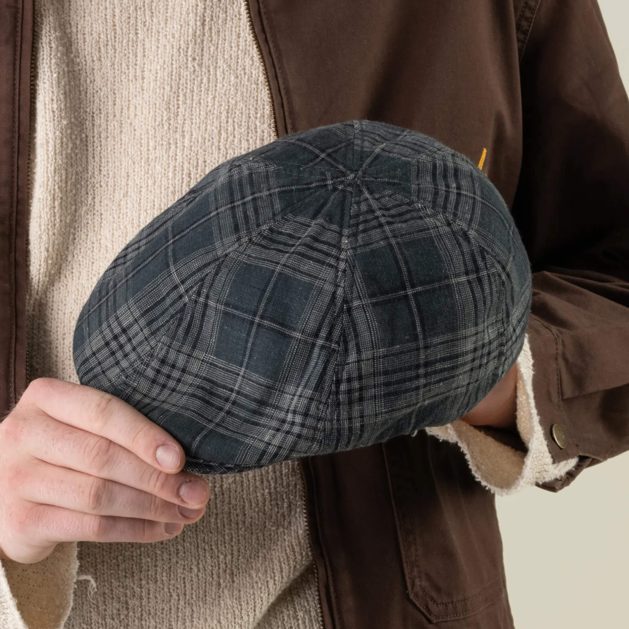 A person wearing a brown jacket and cream sweater holds the Goorin Bros. Glenwood Grove flat cap—crafted from 100% cotton with a headwear chassis in gray plaid—in their left hand.