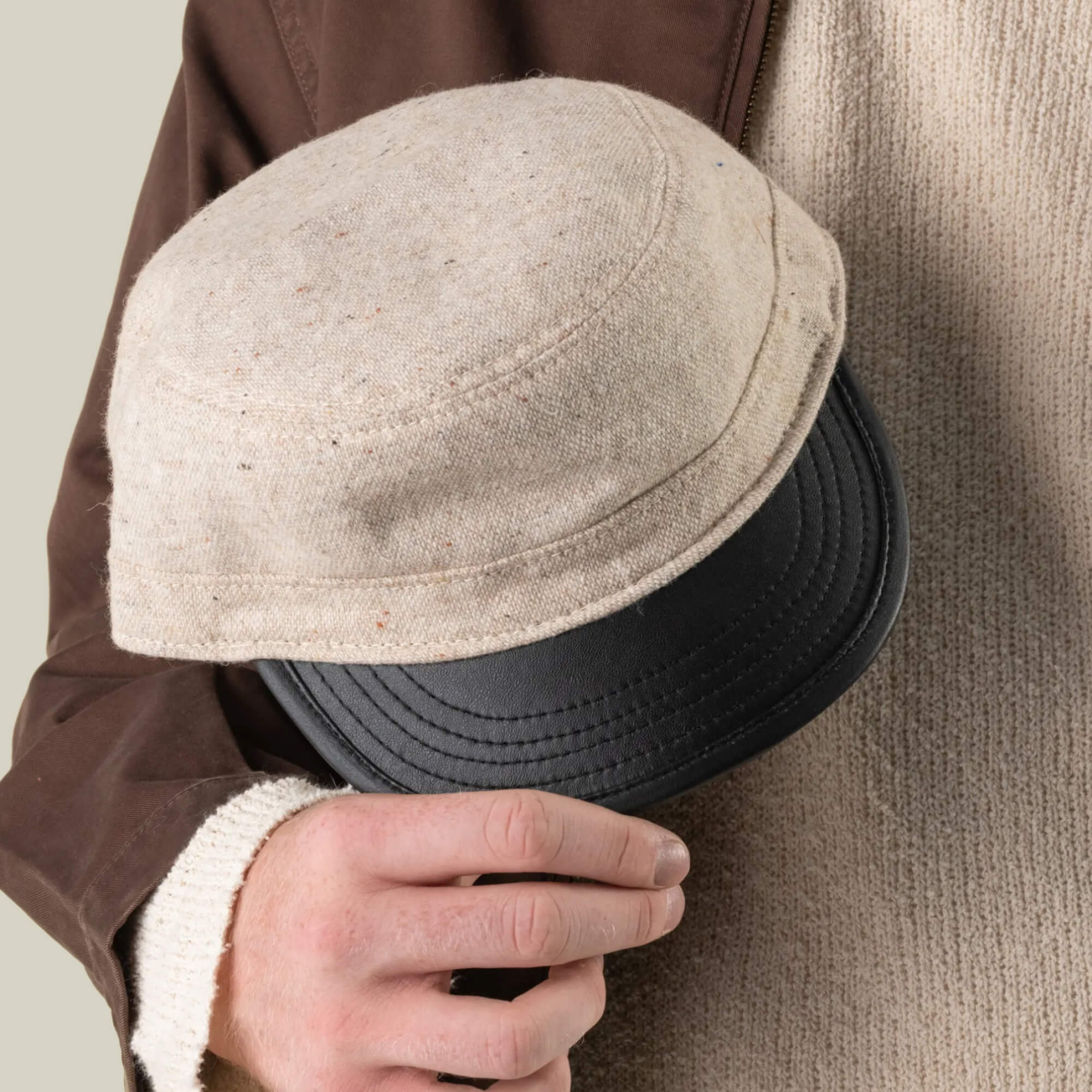 A heritage-minded person wearing a beige sweater and brown jacket holds the Goorin Bros. South Boulder cadet cap—hand cut and sewn in the USA—in their right hand, with the cap partially covering their chest.