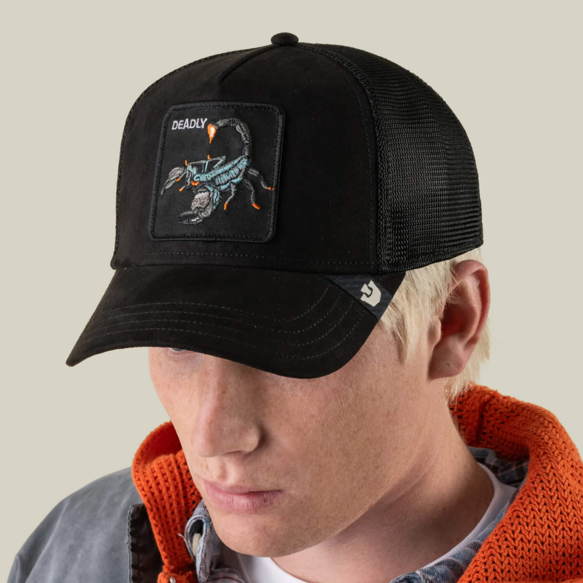 A person wearing the Goorin Bros. Suede Scorpion black trucker hat with a suede front, breathable mesh, and a DEADLY scorpion patch, styled with an orange hoodie and gray jacket, looking down.