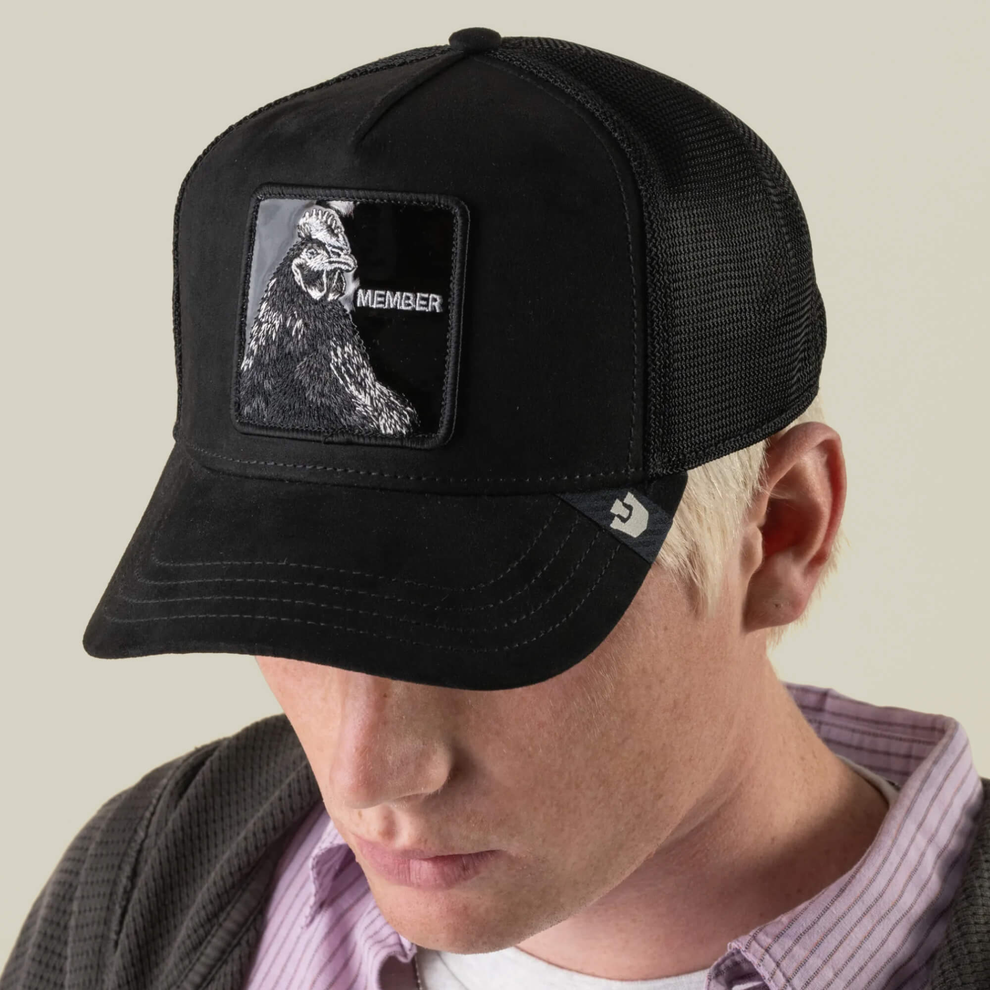 A person wearing a black mesh baseball cap featuring a MEMBER patch with a gorilla design from Goorin Bros., paired with jet-black Black Out Rooster slides, looking down.