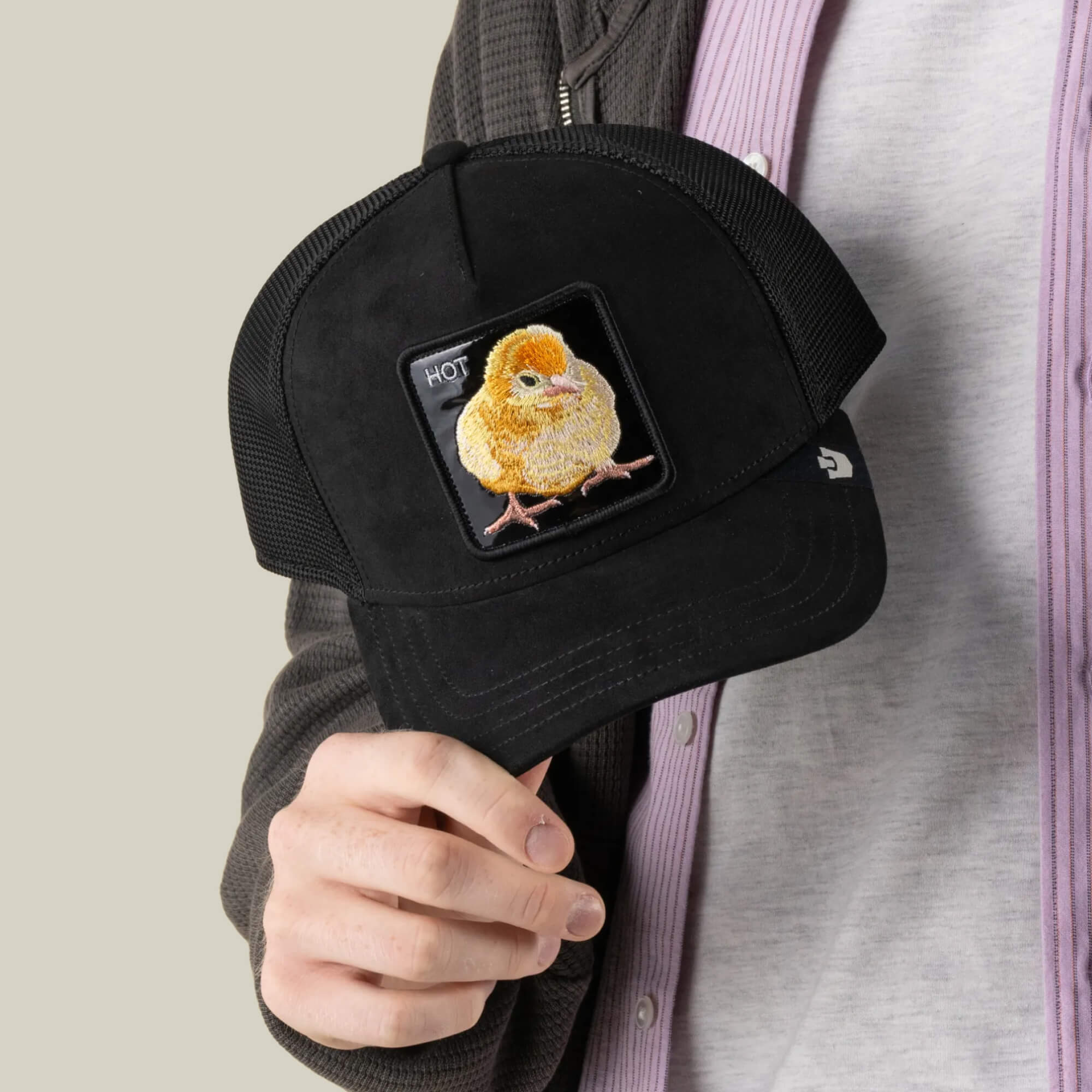 A person holds the Black Out Chick baseball cap by Goorin Bros., featuring a yellow chick patch and the word “HOT” in the upper left corner, designed in a classic Goorin Bros. trucker style.