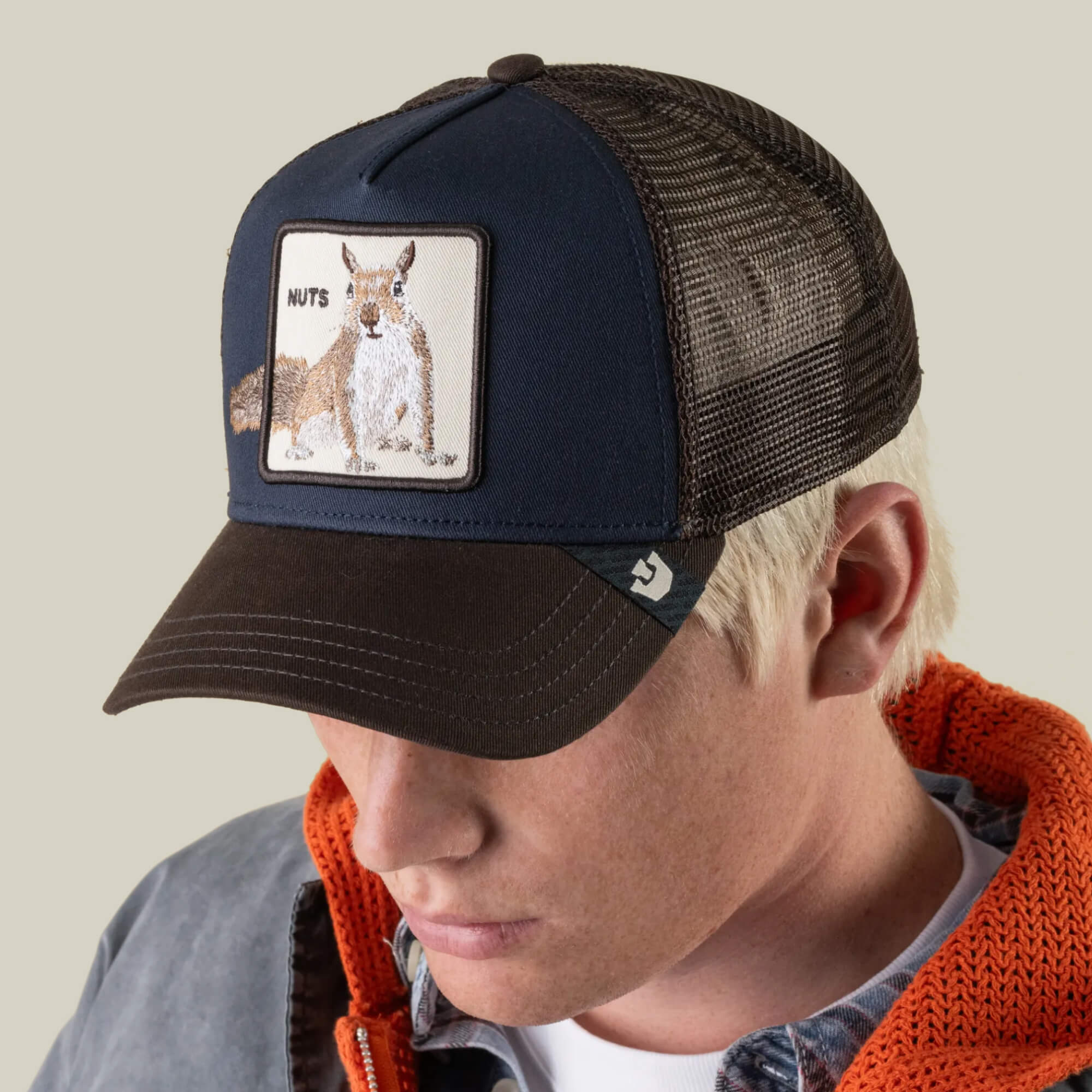 A person wearing the Goorin Bros. “Over the Line Squirrel” black and brown mesh hat, featuring an embroidered patch with a squirrel and the word “NUTS,” paired with a gray jacket and orange hoodie, looking down.