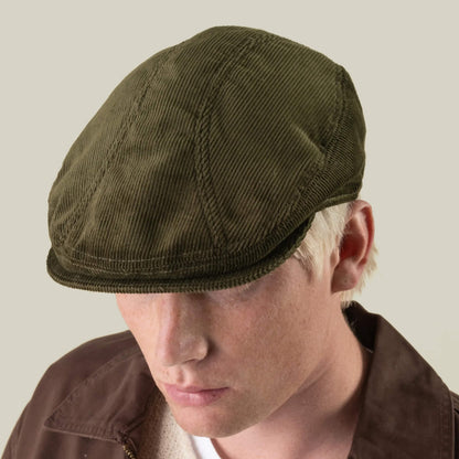 A person wearing the Maxwell Park by Goorin Bros.—a durable, expertly hand-cut olive green corduroy flat cap made in the USA—paired with a brown jacket, facing slightly downward against a plain background.