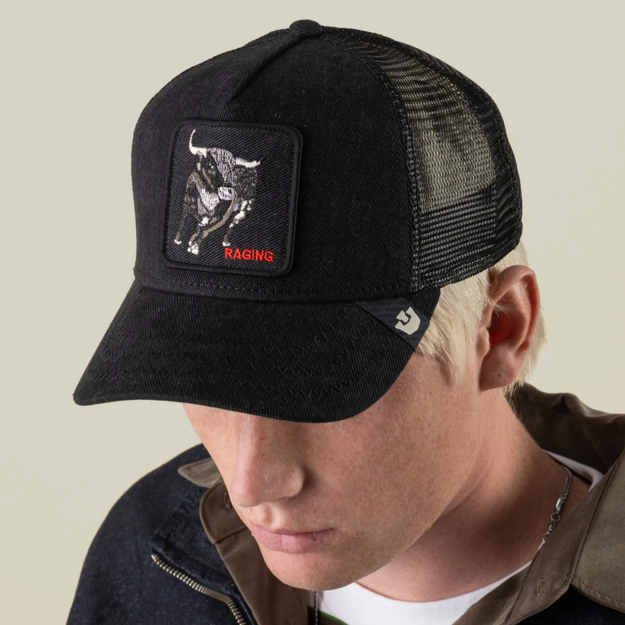 A person wearing the Goorin Bros. Denim Raging trucker hat with a bull patch, paired with a dark jacket and a brown collared shirt, looking down.