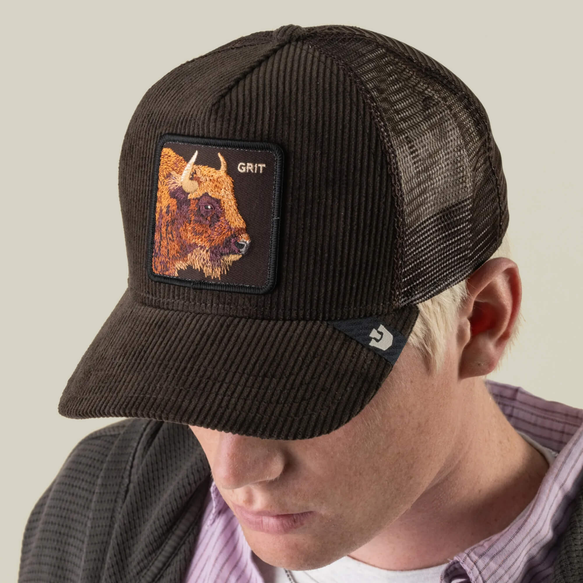 A person wearing the Goorin Bros. Corduroy Grit cap—a dark brown corduroy hat with a bison patch, the word “GRIT,” breathable mesh panels, and a curved brim—looks downward.