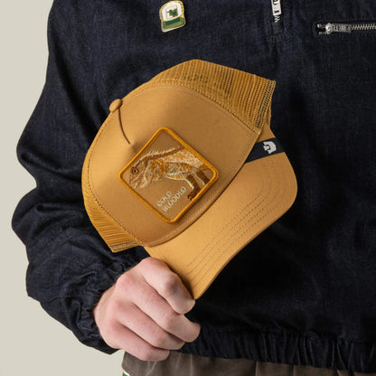 A person in a dark jacket holds a yellow mesh trucker hat from Goorin Bros., featuring a dinosaur patch labeled “SOLID SAURO” on the front; this playful hat is called “Circle of Life - Cold Blooded.”.