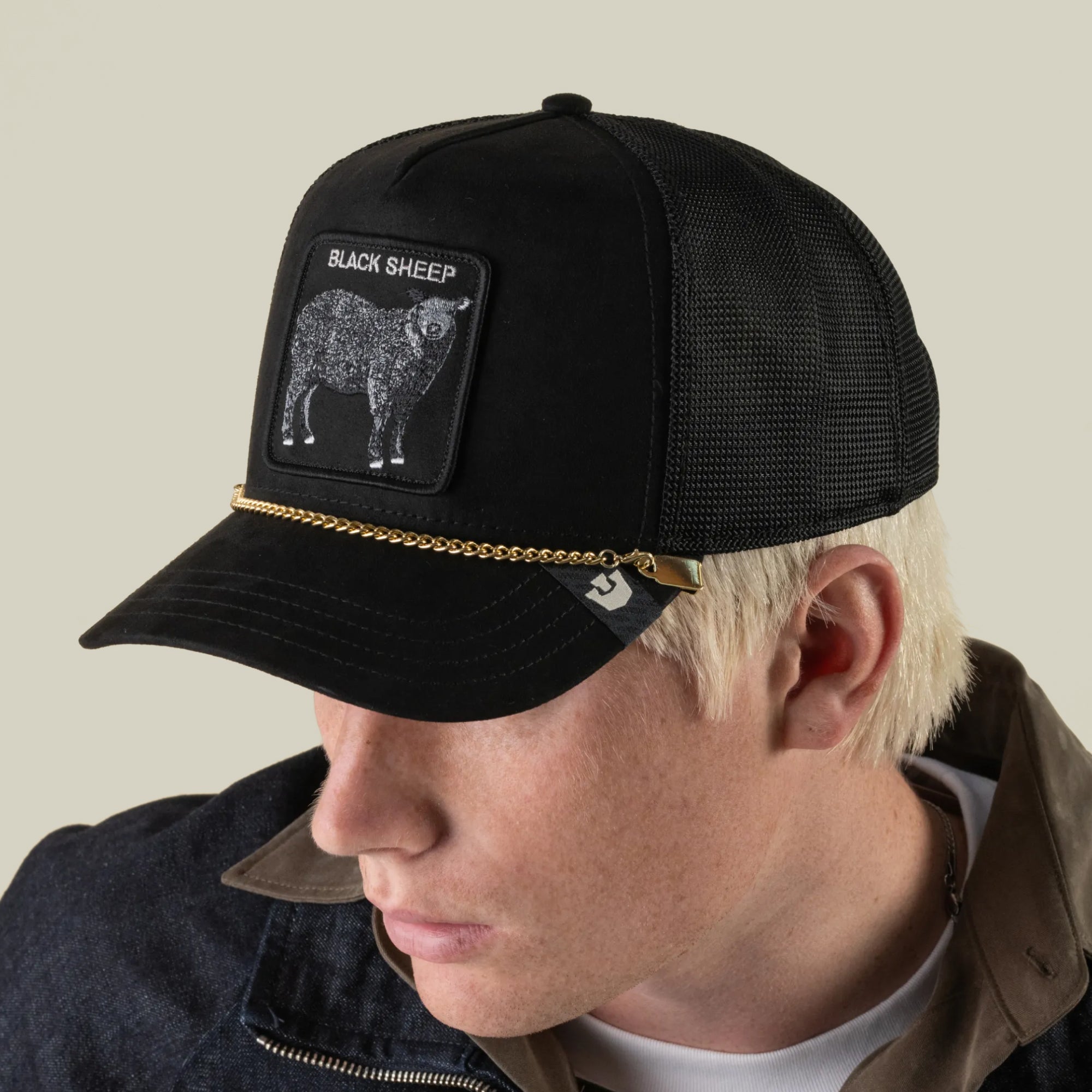 A person wearing a black mesh trucker hat by Goorin Bros. featuring a Black Sheep patch and gold detailing, accessorized with the Mini CapLoc Chain as stylish hat jewelry, looks down and to the side against a plain background.