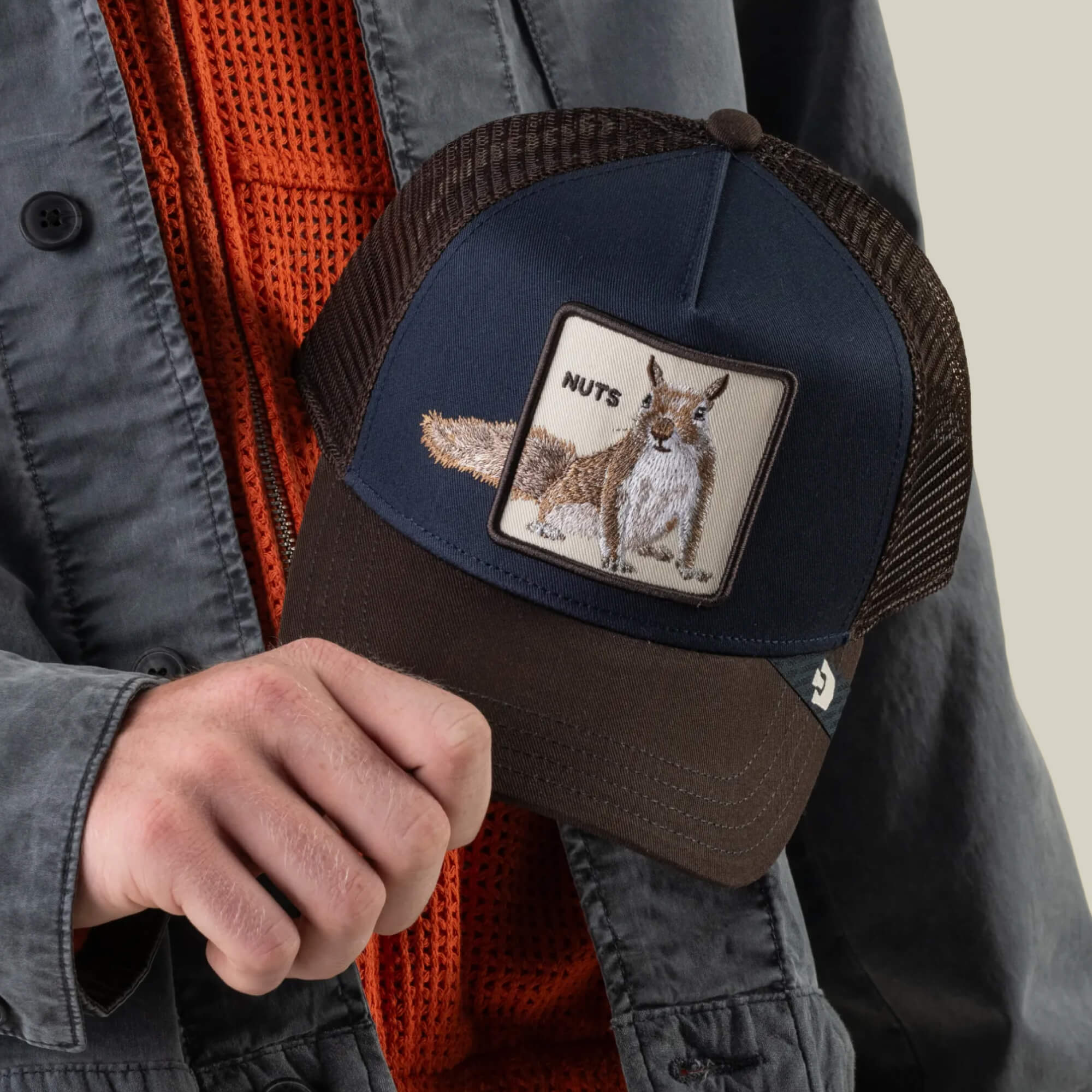 A person holds the Goorin Bros. “Over the Line Squirrel” hat in brown and navy, featuring an embroidered patch with a squirrel and the word “NUTS,” designed with an adjustable fit for comfort.