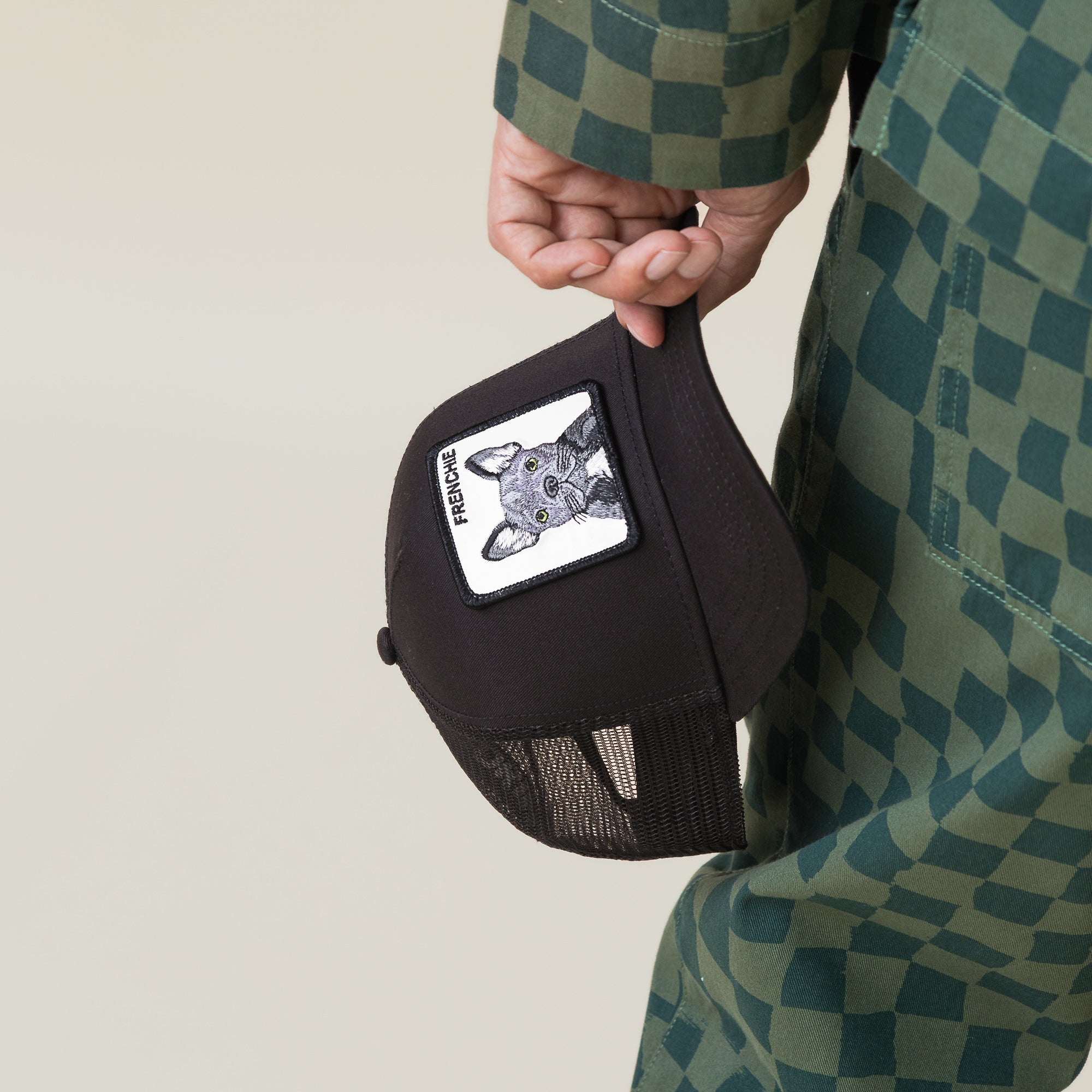 Someone holding the Goorin Bros. Frenchie Trucker—a cute black cap featuring a dog face patch and the word "FRENCHIE"—while dressed in a green checkered outfit.