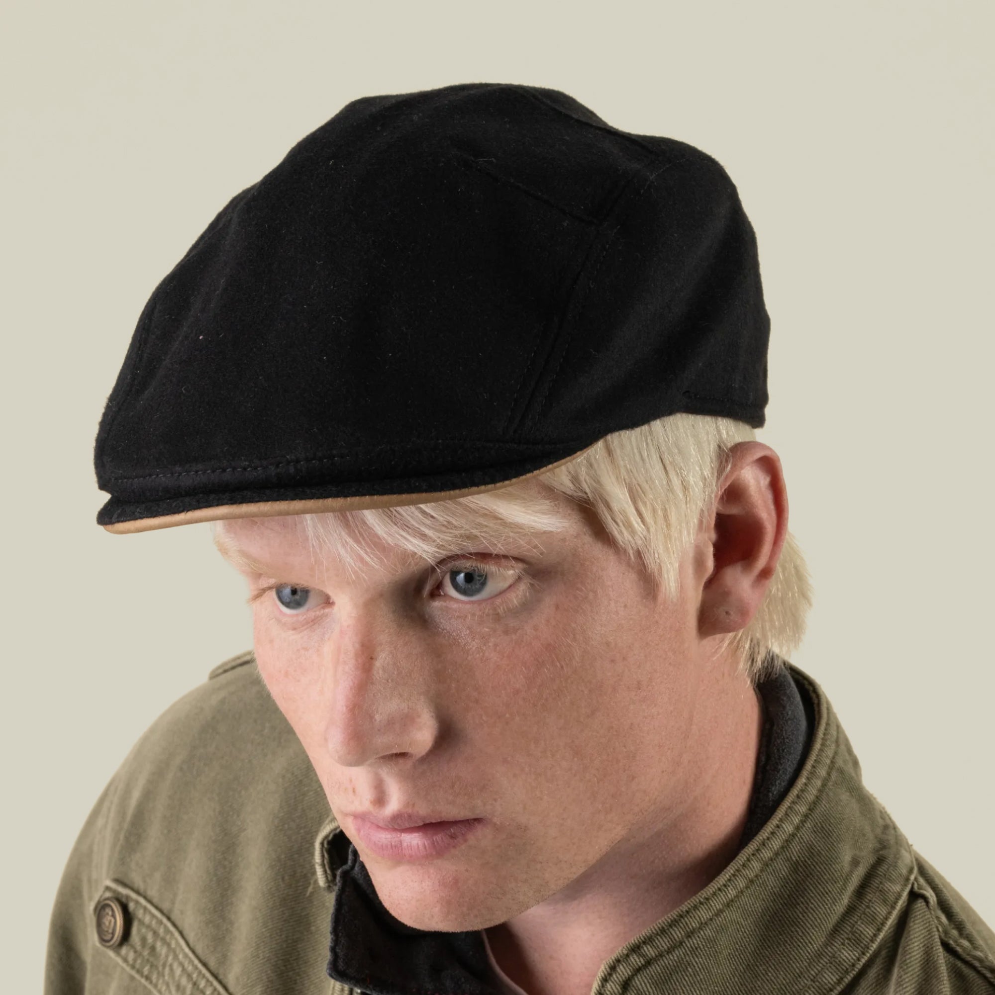 A person with light blond hair wearing the Goorin Bros. Cornerstone handmade black flat cap and an olive green jacket, looking slightly downward against a plain background.